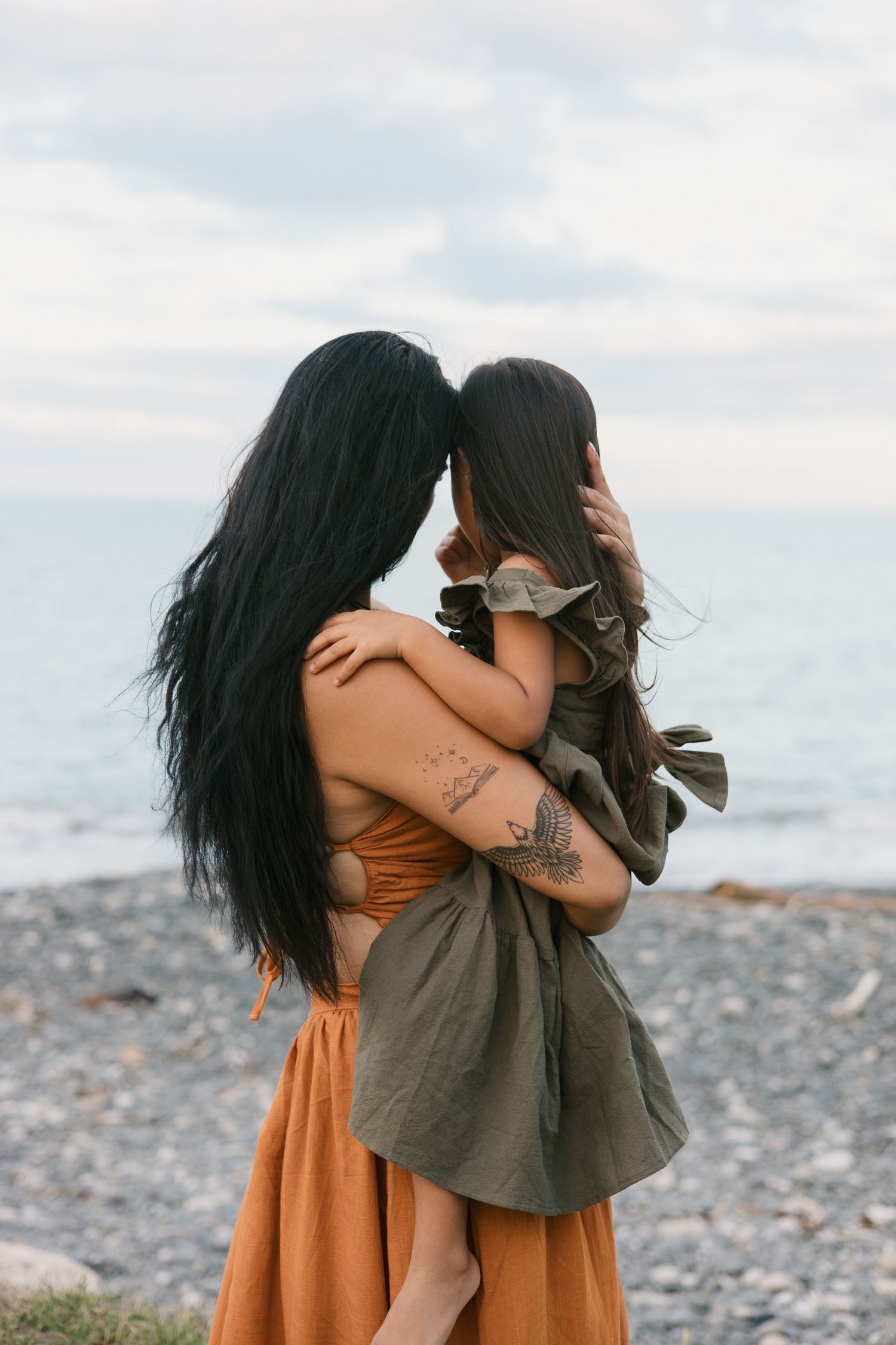 Mother and daughter share an intimate embrace on a rocky beach with ocean waves in the background.