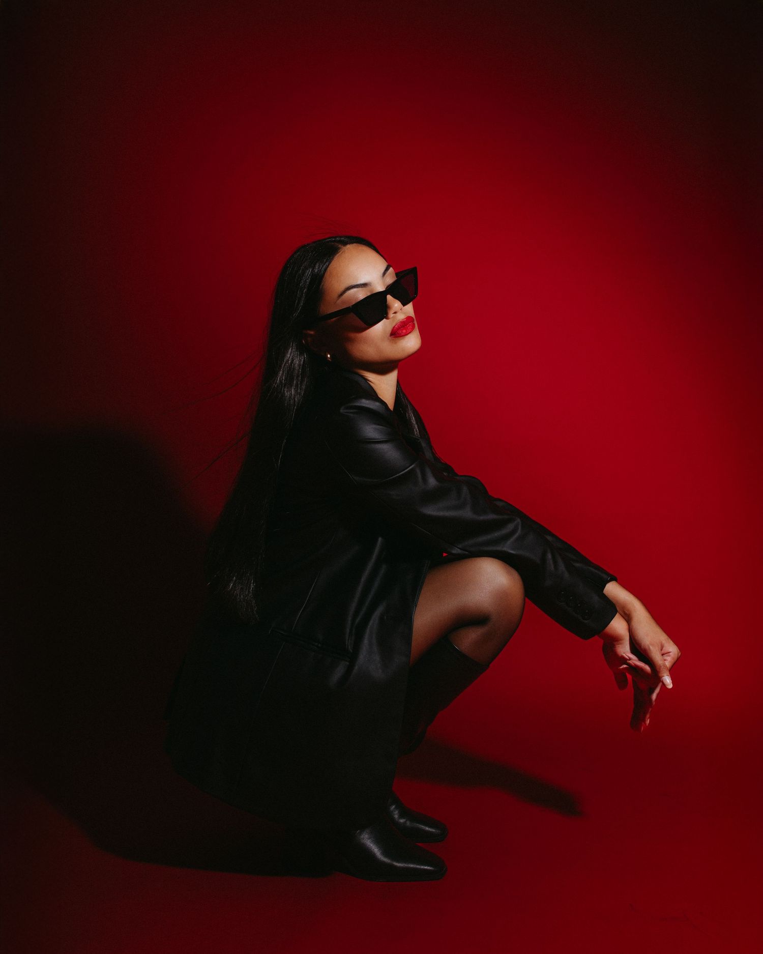Dramatic fashion portrait against red backdrop featuring dark outfit with sunglasses in crouching pose.