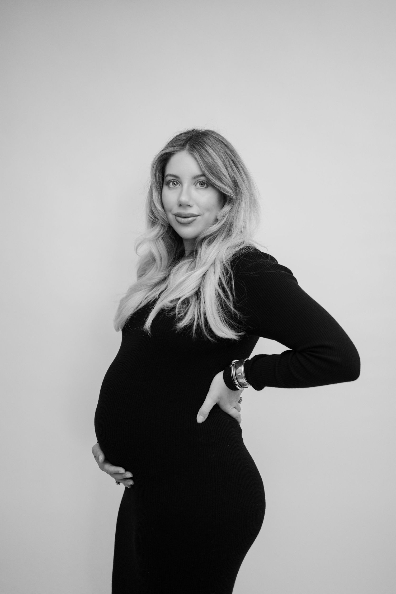 Black and white maternity photo showing a pregnant woman in a black dress posing against a light background.