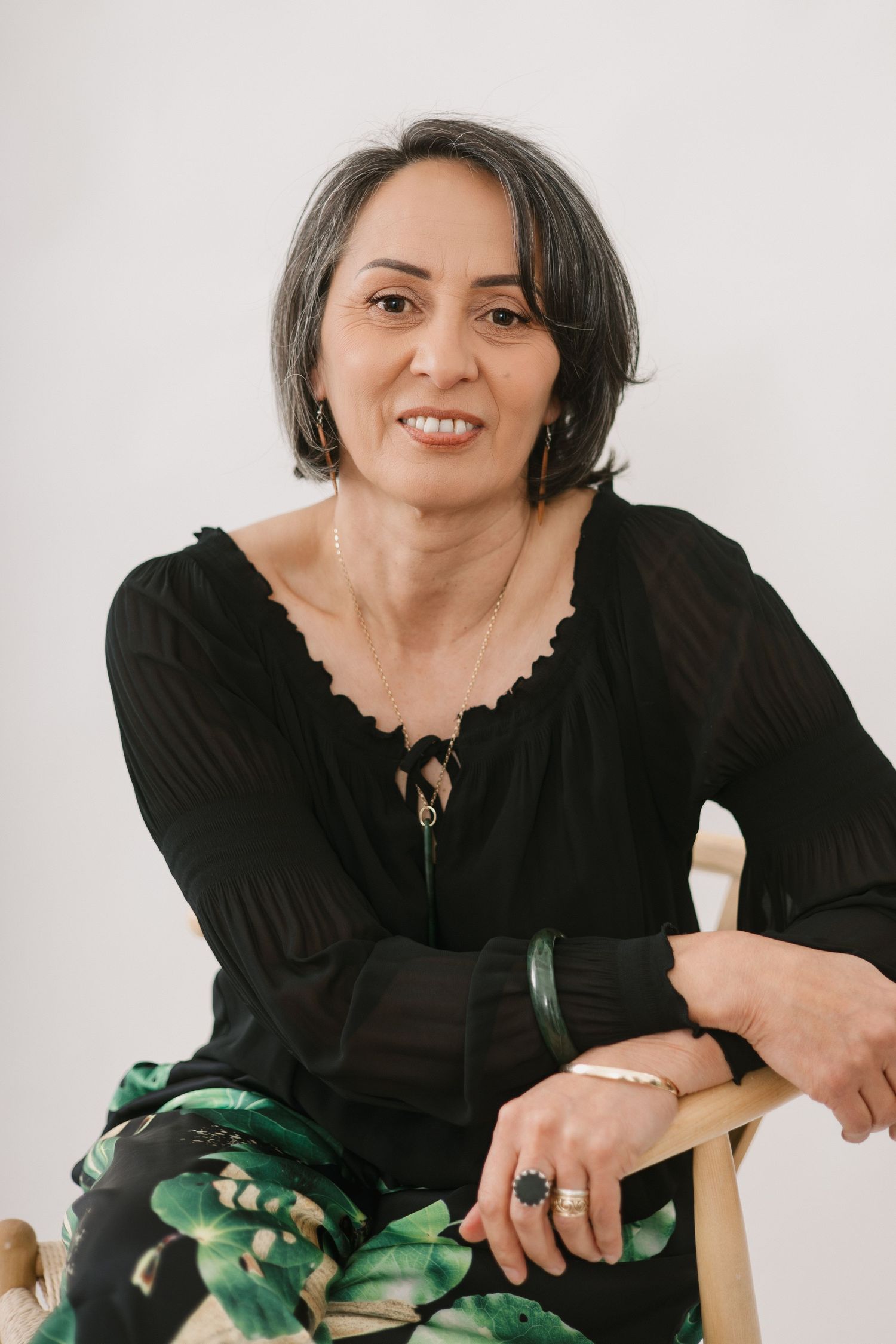 Woman in black top with floral print pants sits casually on wooden chair against white backdrop.