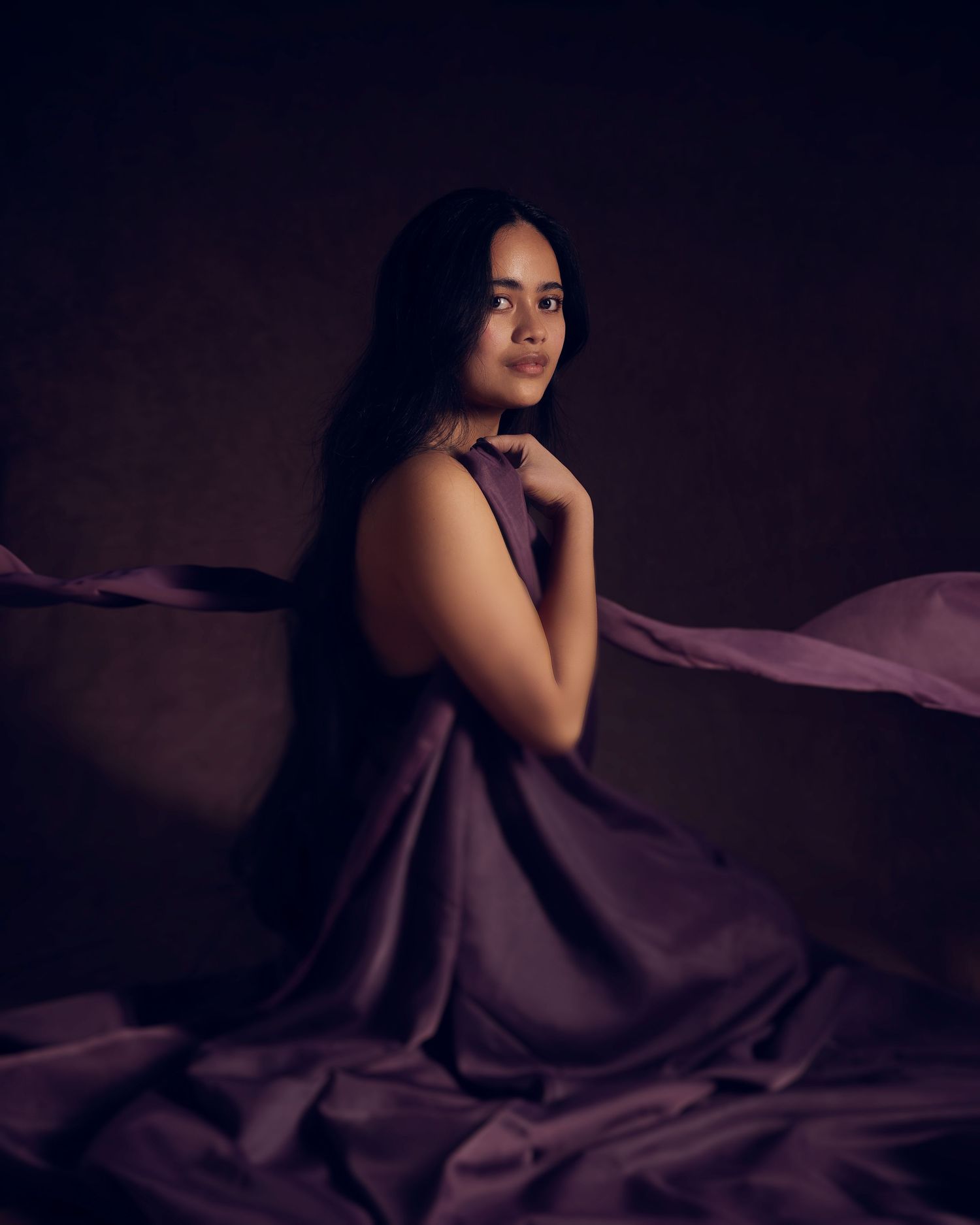 Artistic portrait with soft lighting shows figure wrapped in flowing purple fabric against dark moody background.