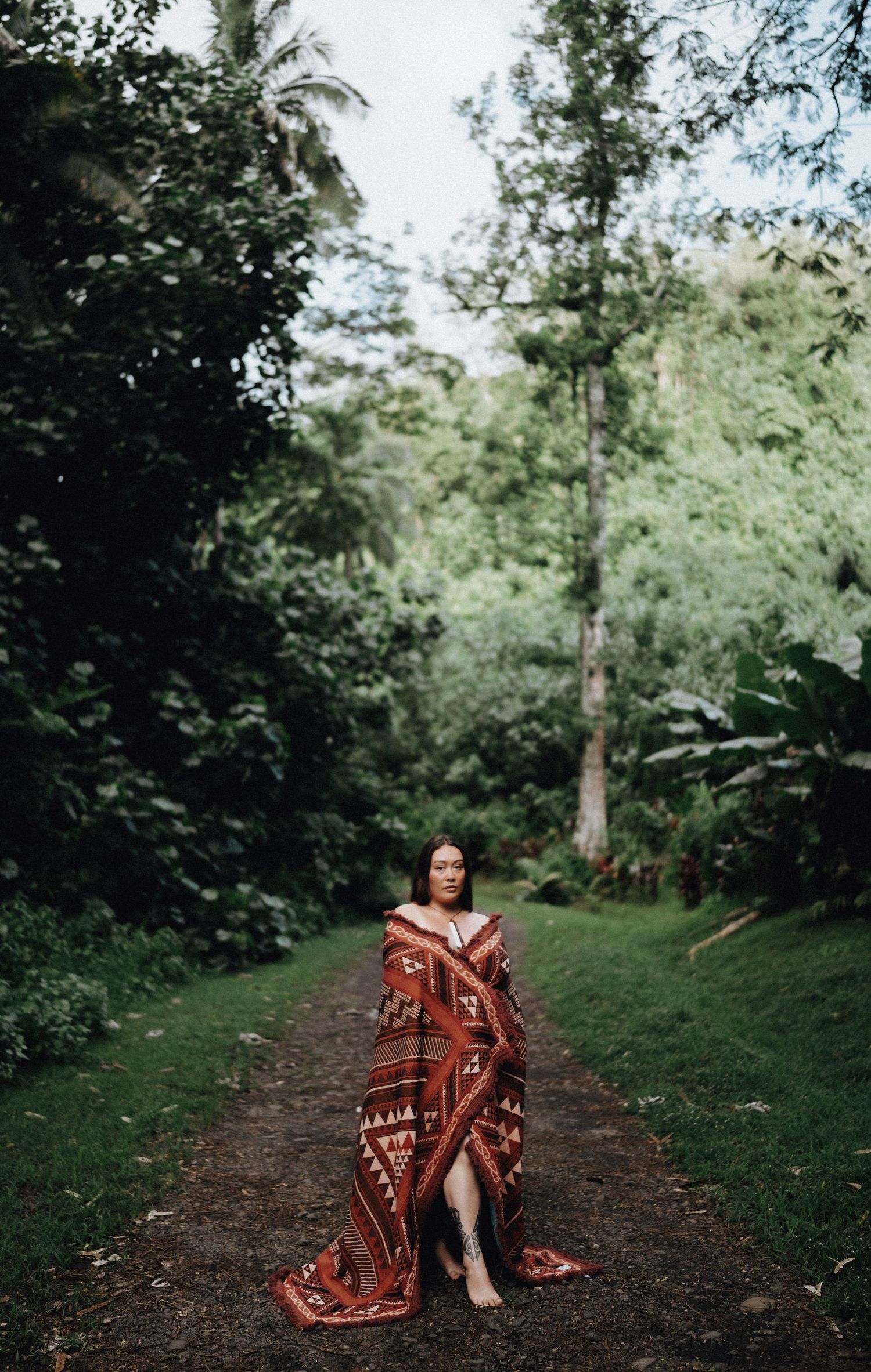 Person wrapped in patterned blanket standing on forest path surrounded by lush greenery.