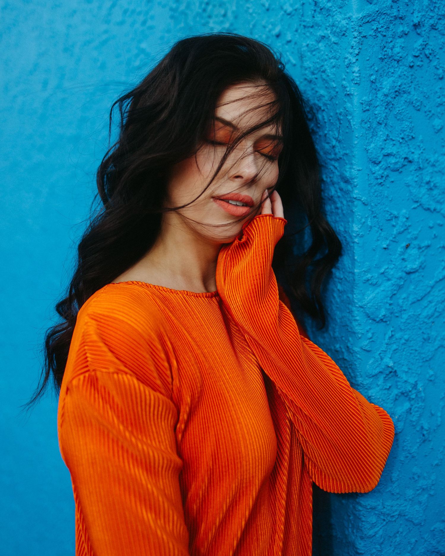 Person with flowing dark hair wearing a bright orange sweater poses against a vivid blue wall background.
