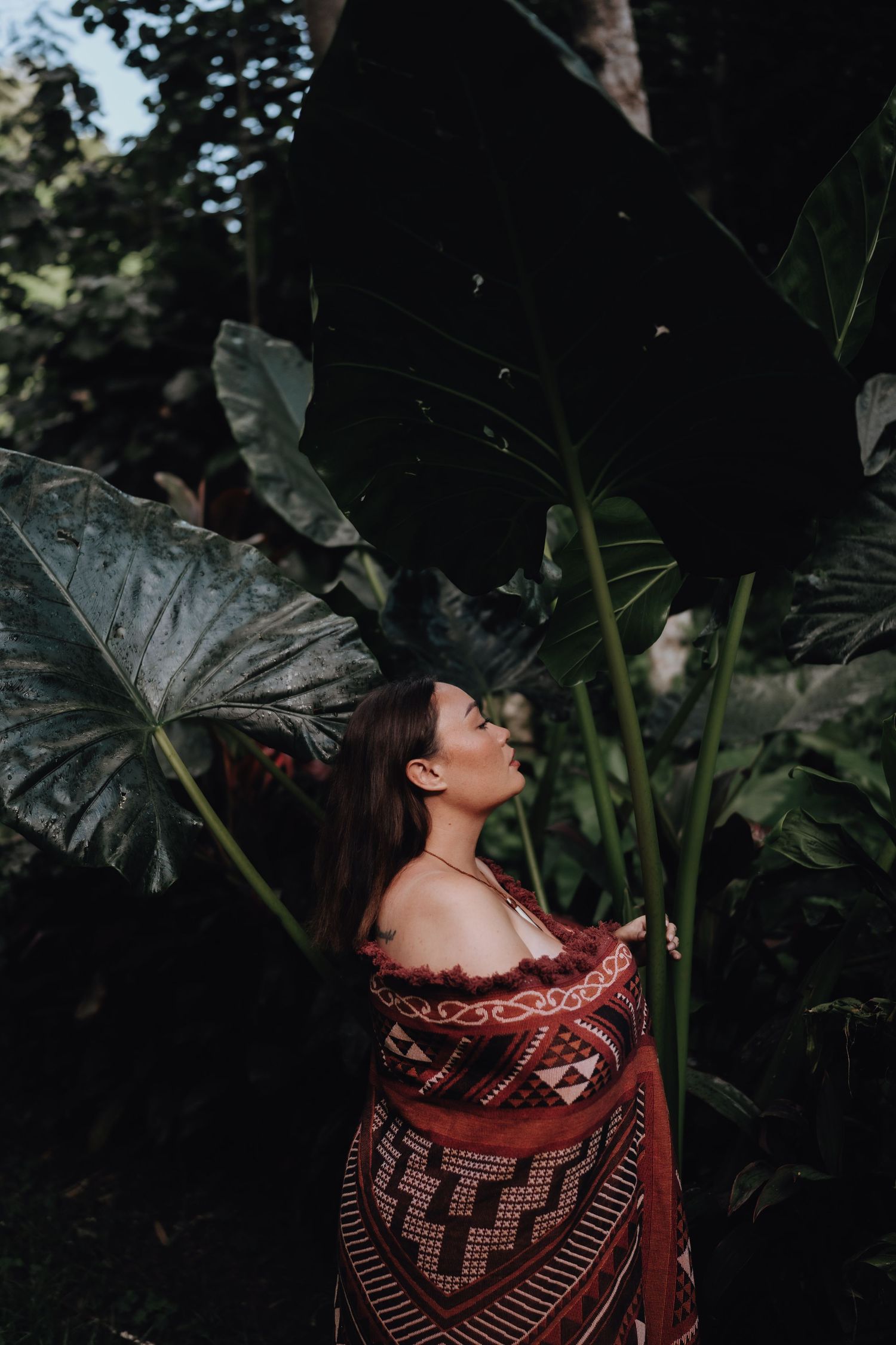 A person stands among large tropical leaves wearing a patterned wrap.
