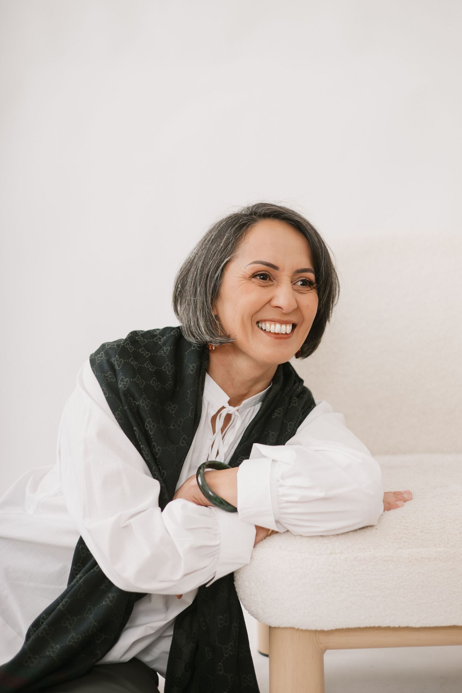 Woman with silver-gray bob haircut smiles while wearing black cardigan and white blouse in minimalist setting.