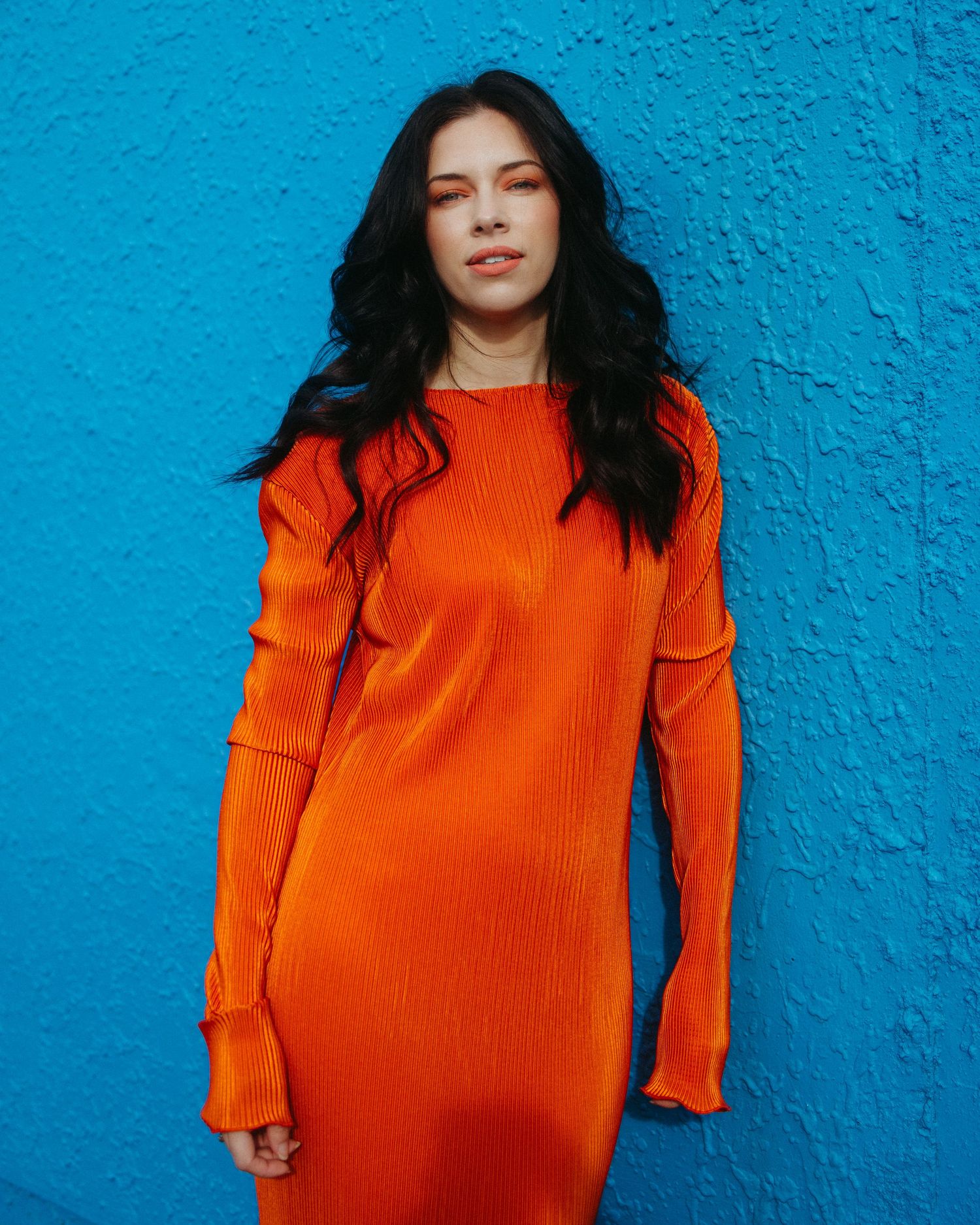A person in a vibrant orange ribbed sweater dress stands against a bright turquoise blue textured wall.