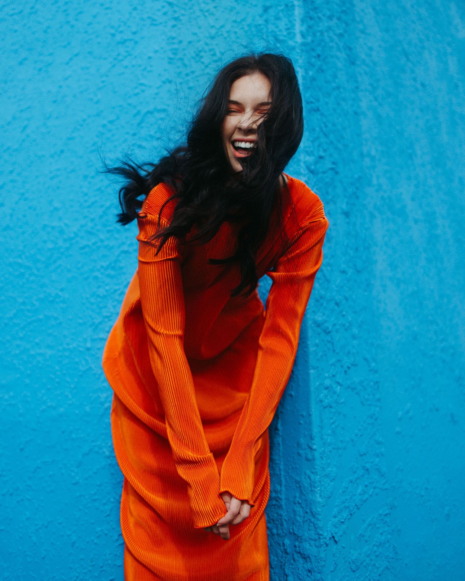 A joyful person in a vibrant orange dress poses expressively against a bright turquoise blue wall with flowing dark hair.