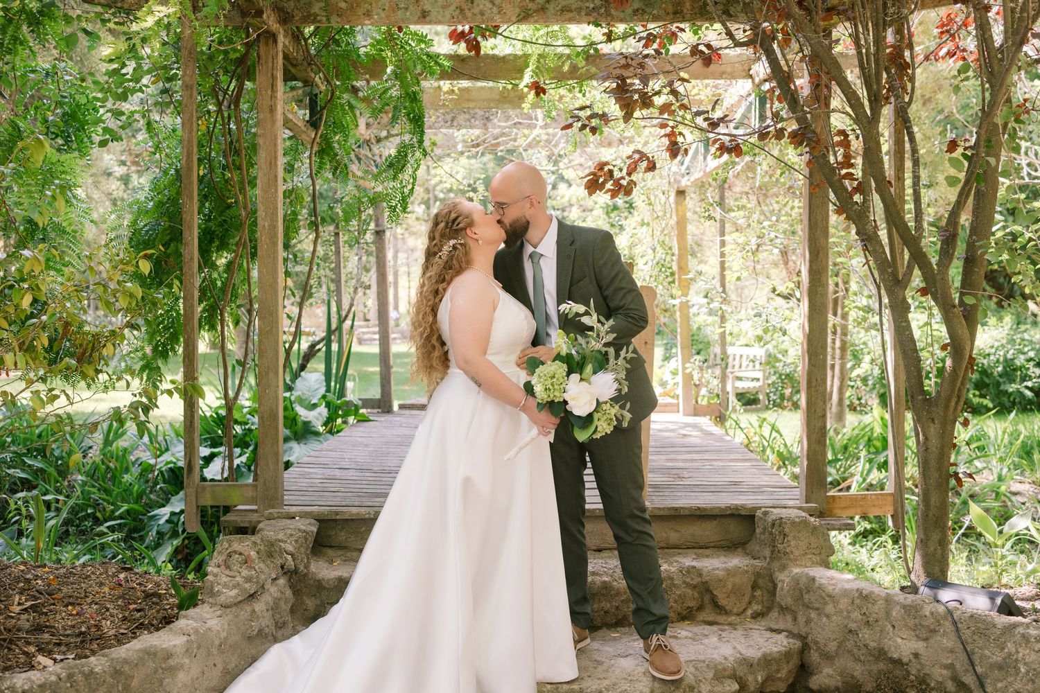 Bride and groom share romantic moment under lush garden pergola with greenery and white flowers.