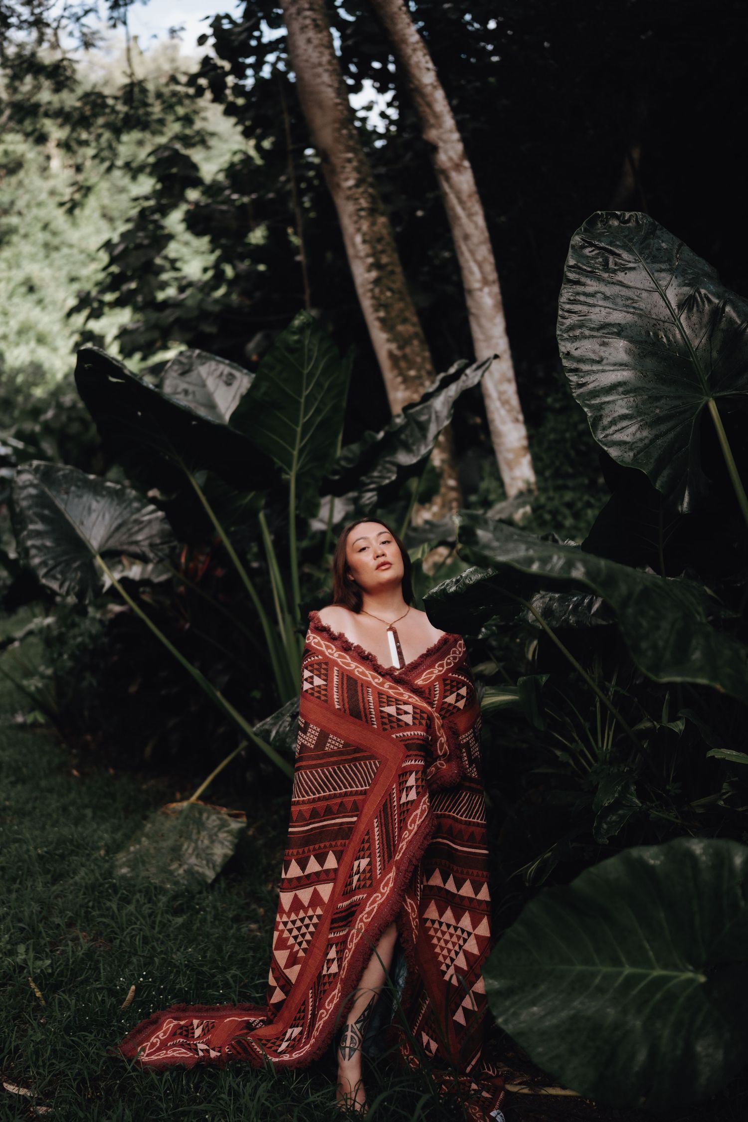 A woman in a red patterned dress poses gracefully against lush tropical foliage in a moody forest setting.