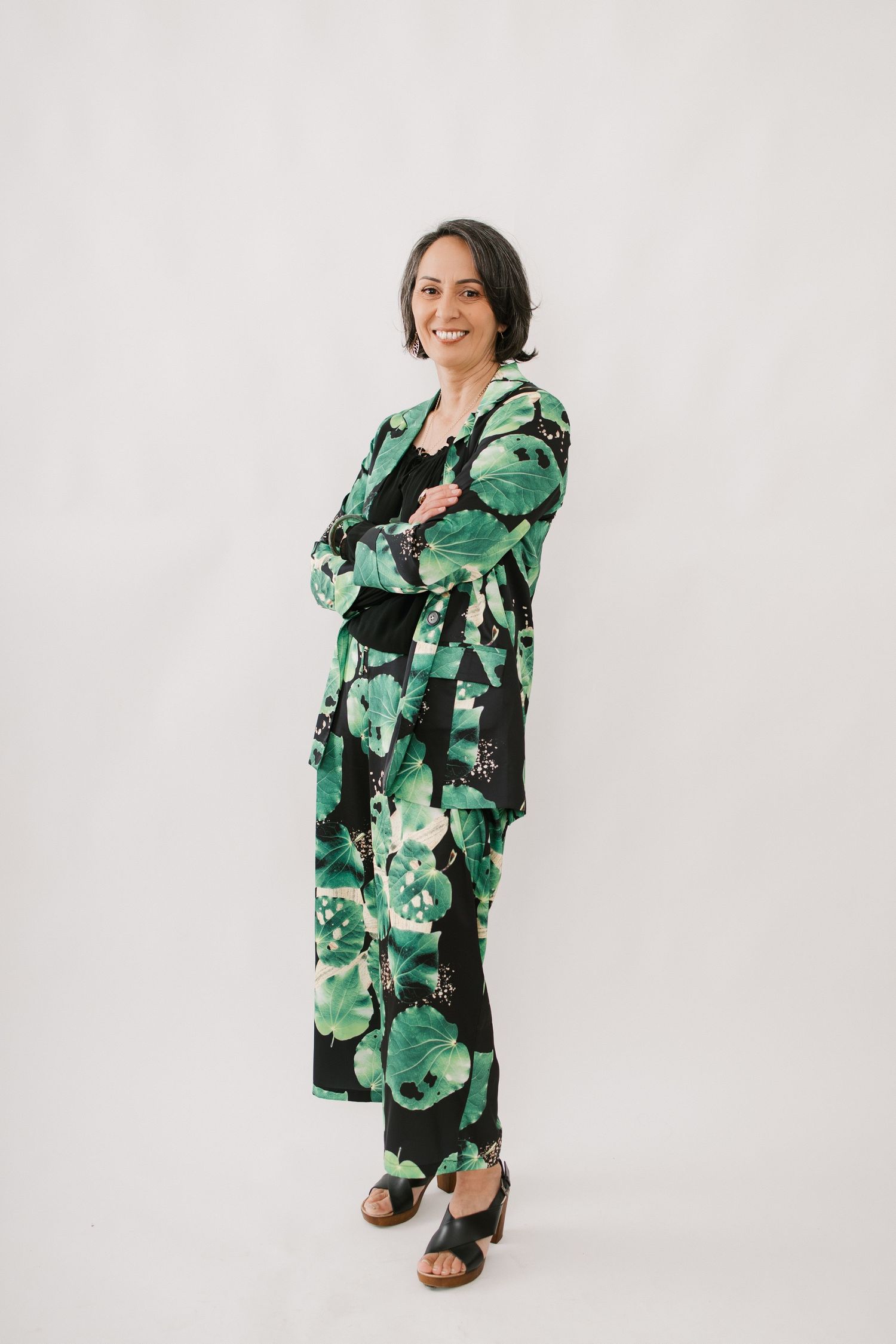 Woman wearing green and black floral print jumpsuit poses against white background with crossed arms.