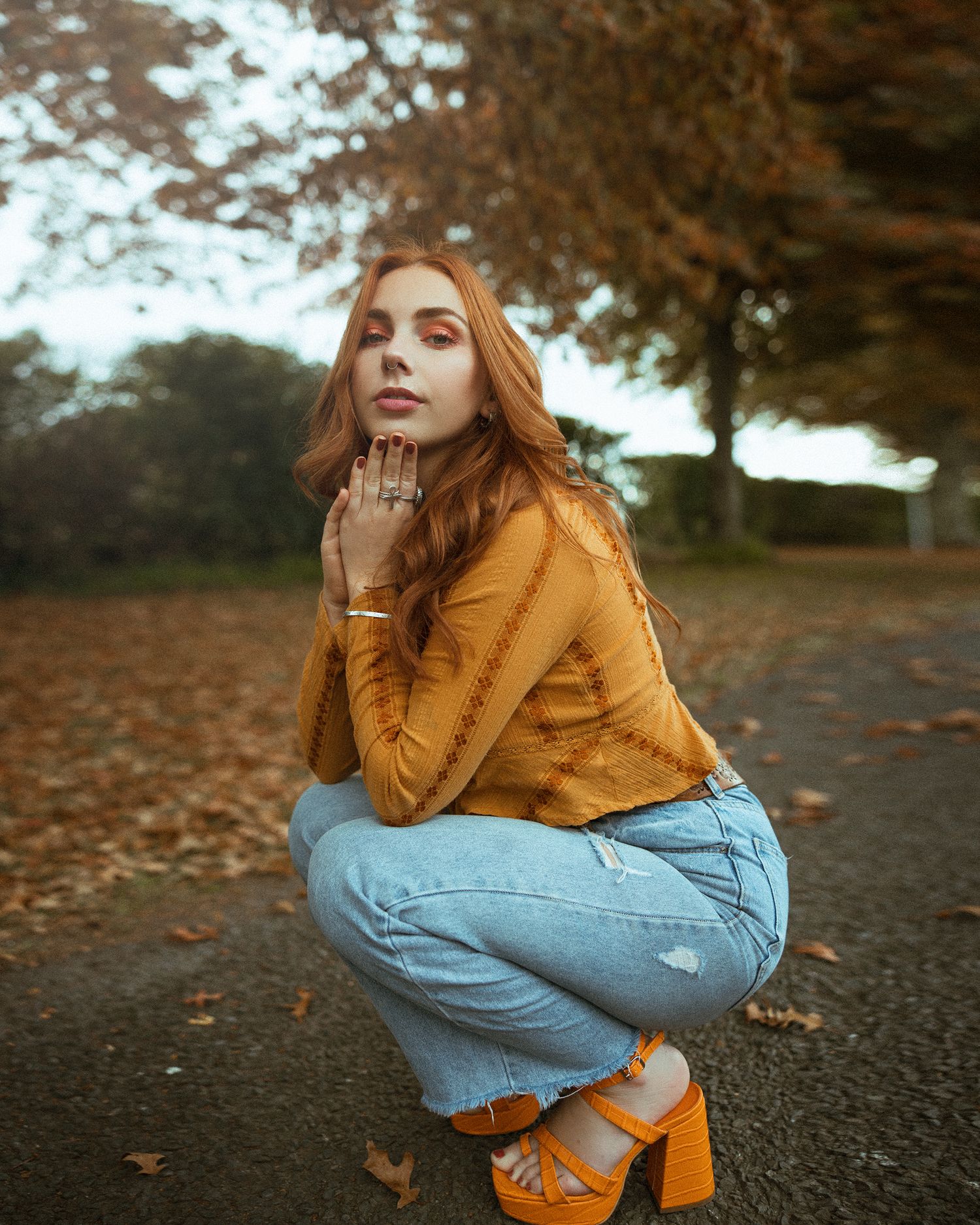 A woman in a mustard sweater and light denim jeans crouches on a path covered in autumn leaves.