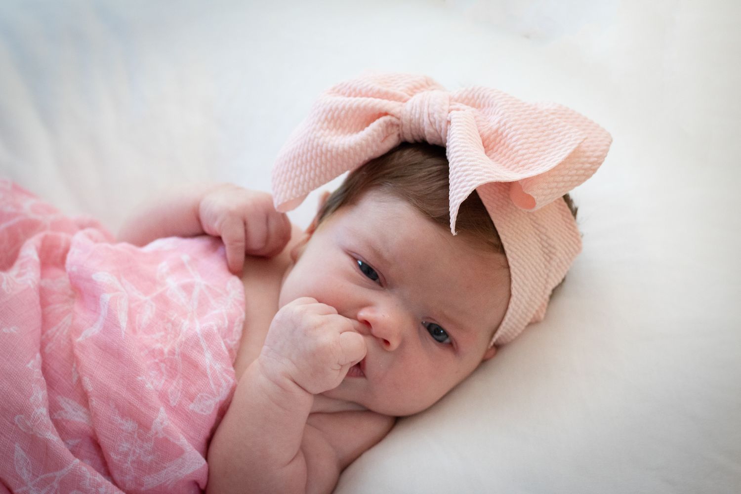 Baby wearing pink bow headband and dress resting on soft white surface.