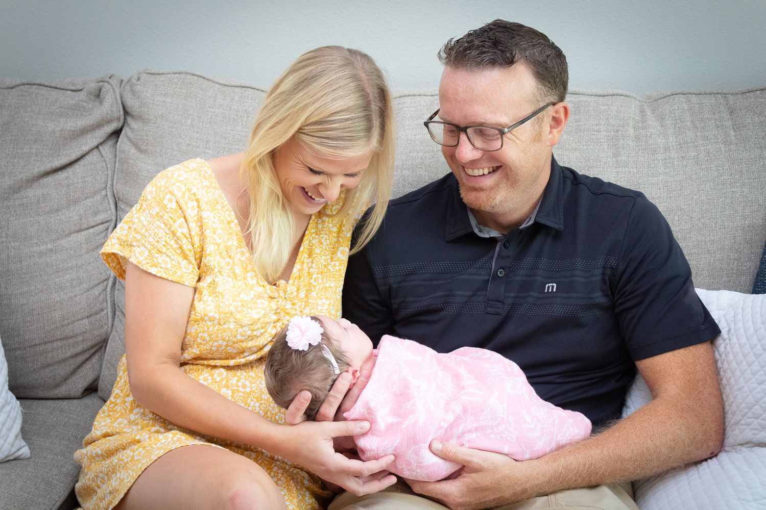 Parents in yellow and navy outfit holding baby in pink on grey couch.