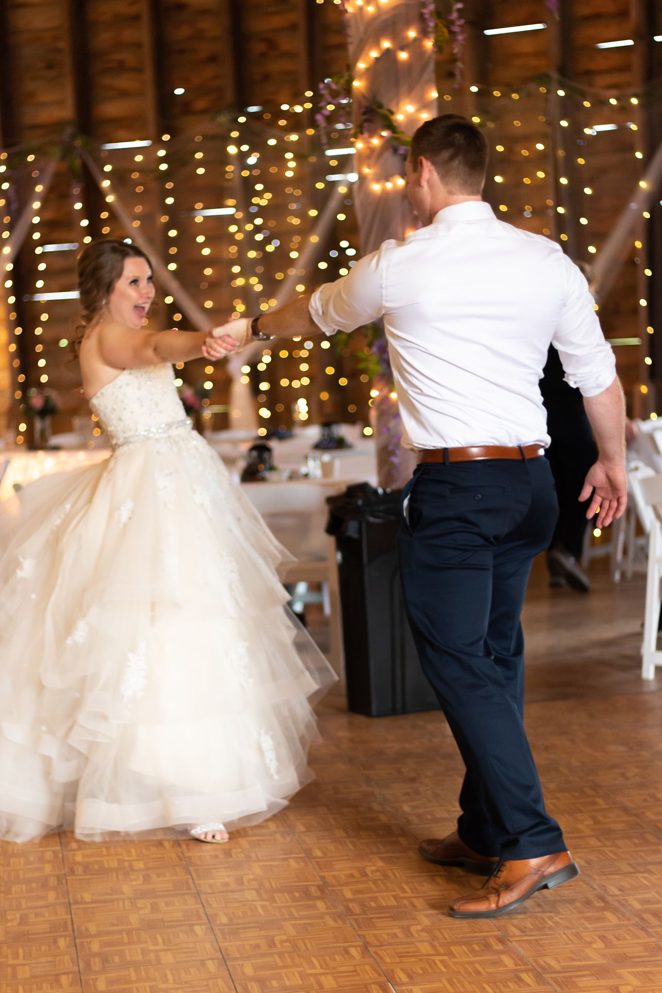 A couple dances in a rustic barn venue decorated with twinkling string lights.