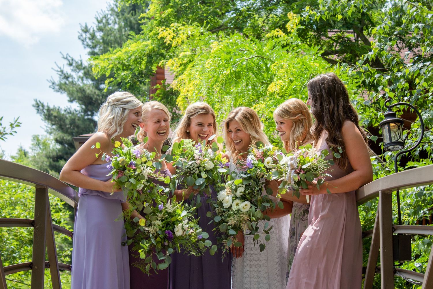 A group in pastel purple and pink bridesmaid dresses gather together looking down at floral bouquets in a garden setting.