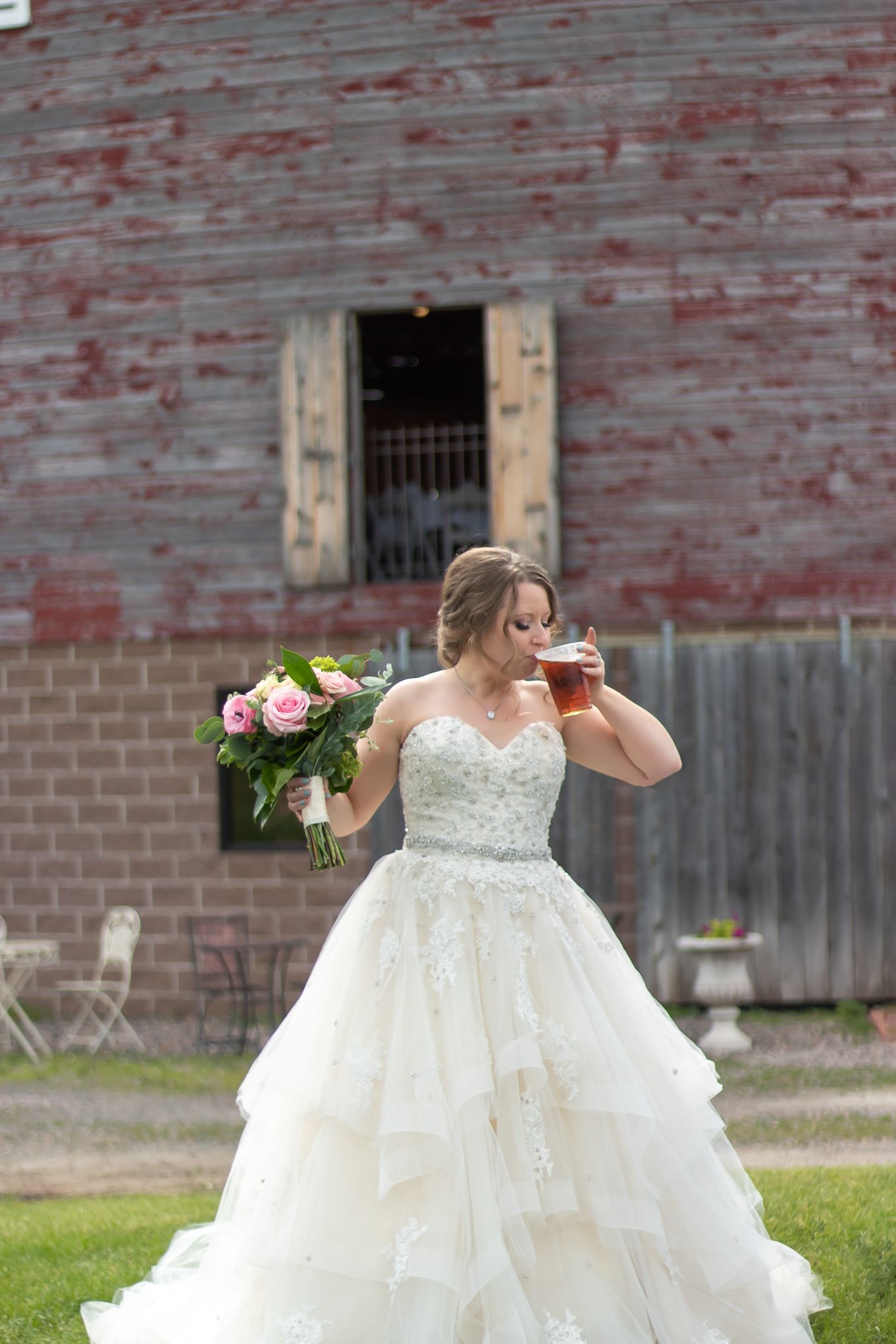 A bride in a layered ballgown holds flowers and a drink near a rustic red barn.