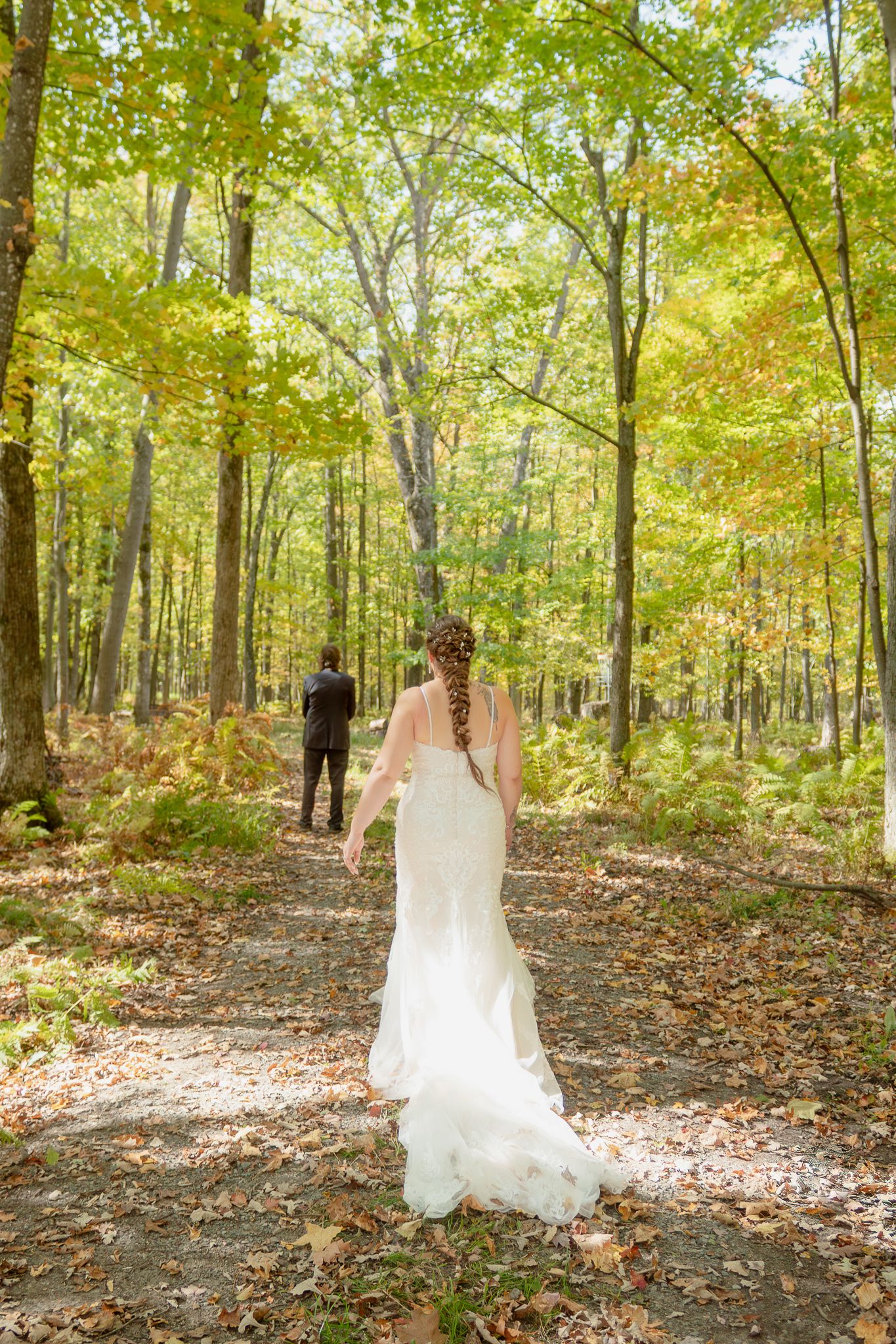 A wedding dress trails on fallen leaves as someone walks through an autumn forest.