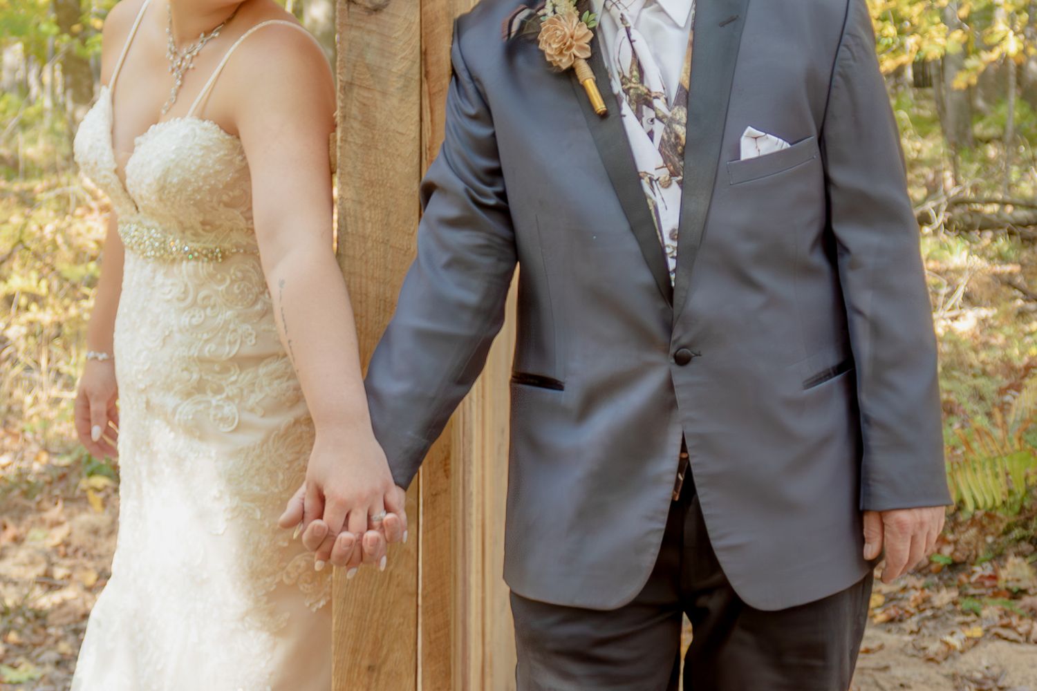 Close up shot of a couple in wedding attire holding hands outdoors, with bride in lace gown and groom in gray suit.
