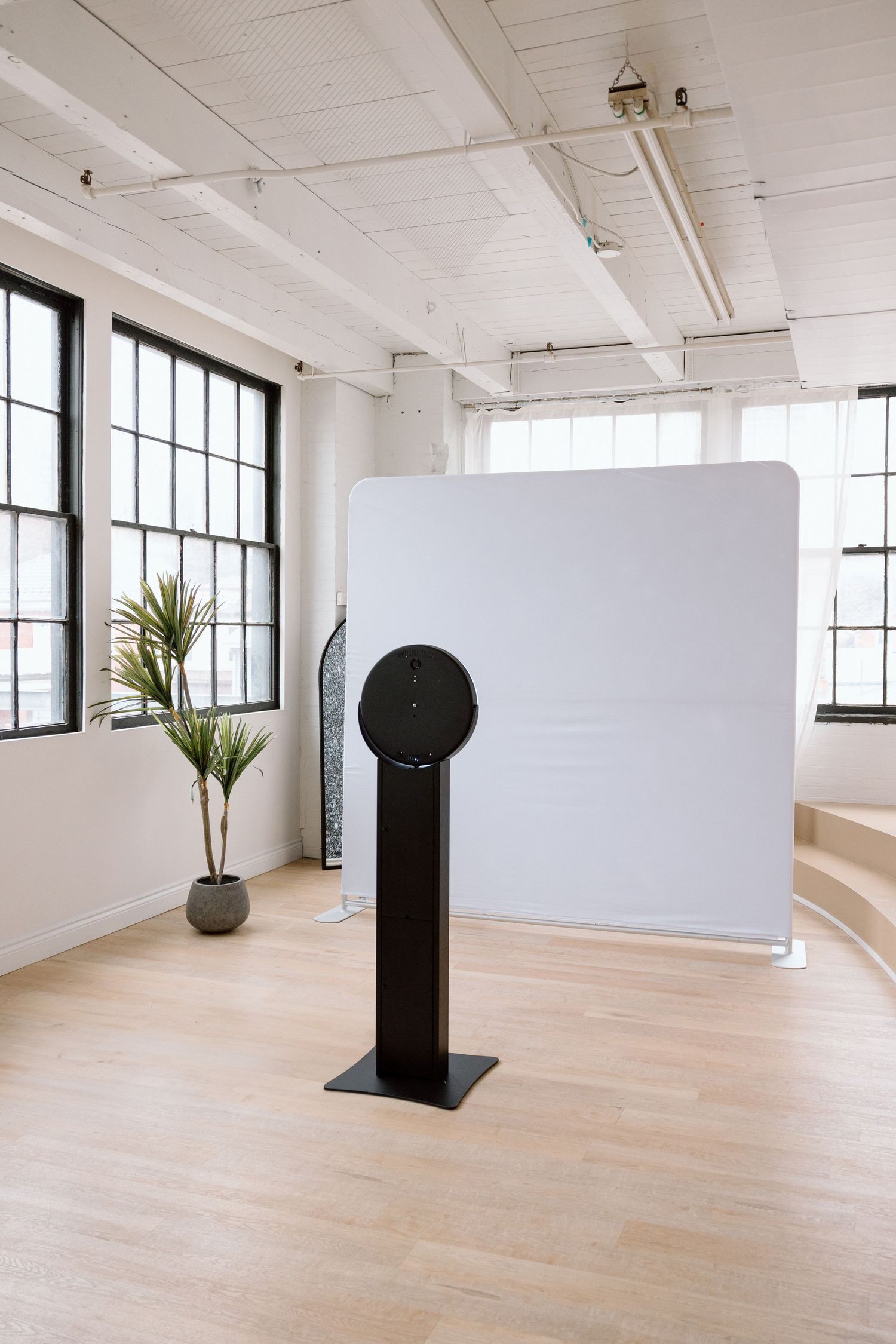 Minimalist photo booth setup with white backdrop and black stand in bright industrial space.