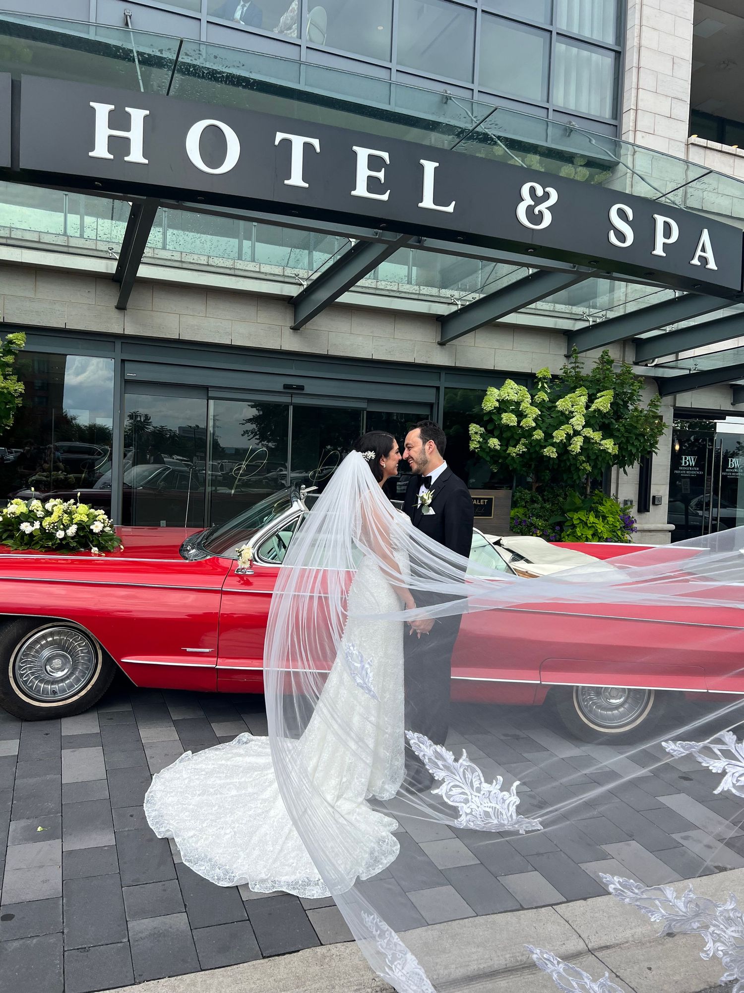 A long white bridal veil flowing in front of a luxury hotel and spa entrance with a red vintage car visible.