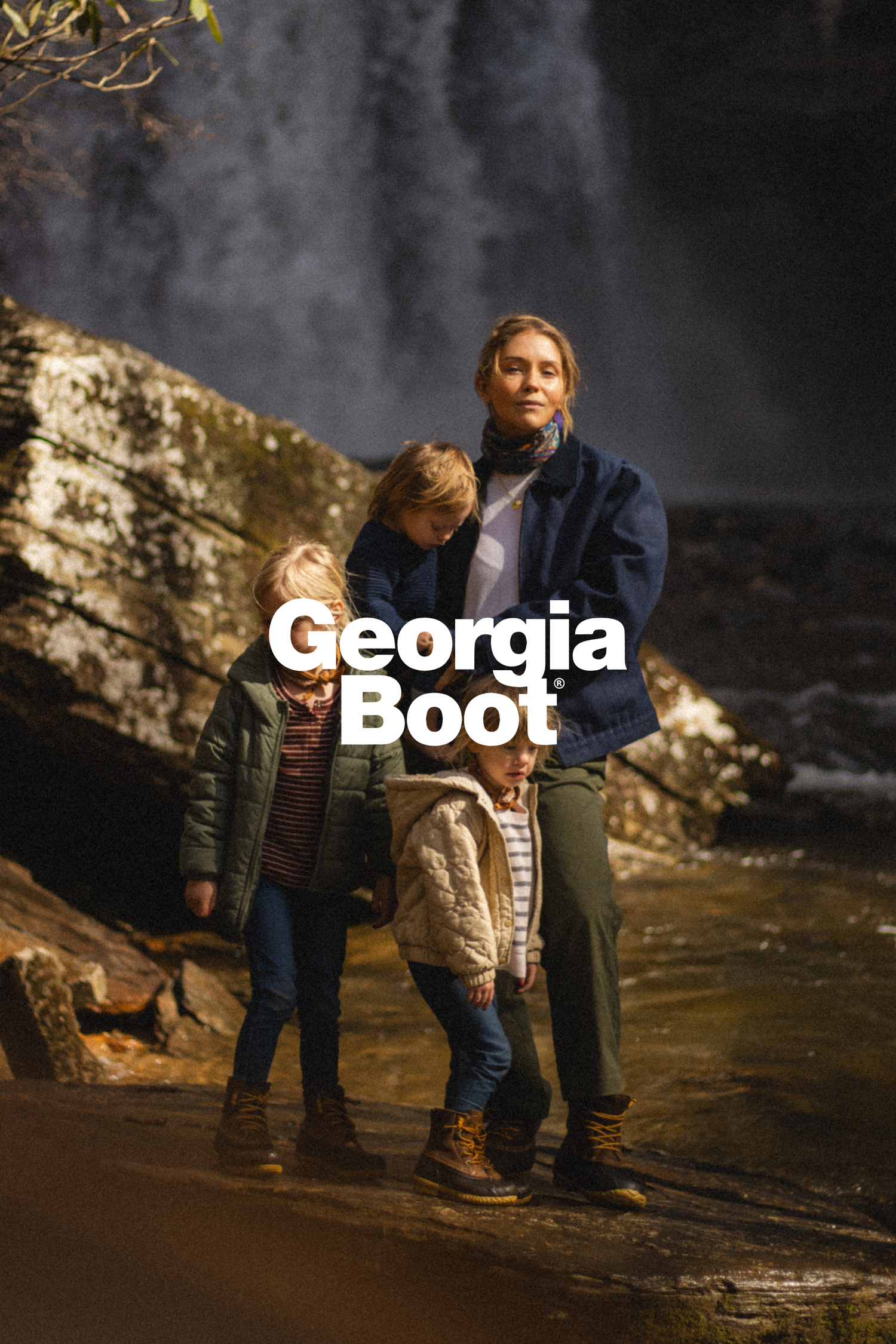 Two people stand on a rocky surface near a misty waterfall with Georgia Boot branding overlaid on the outdoor scene.