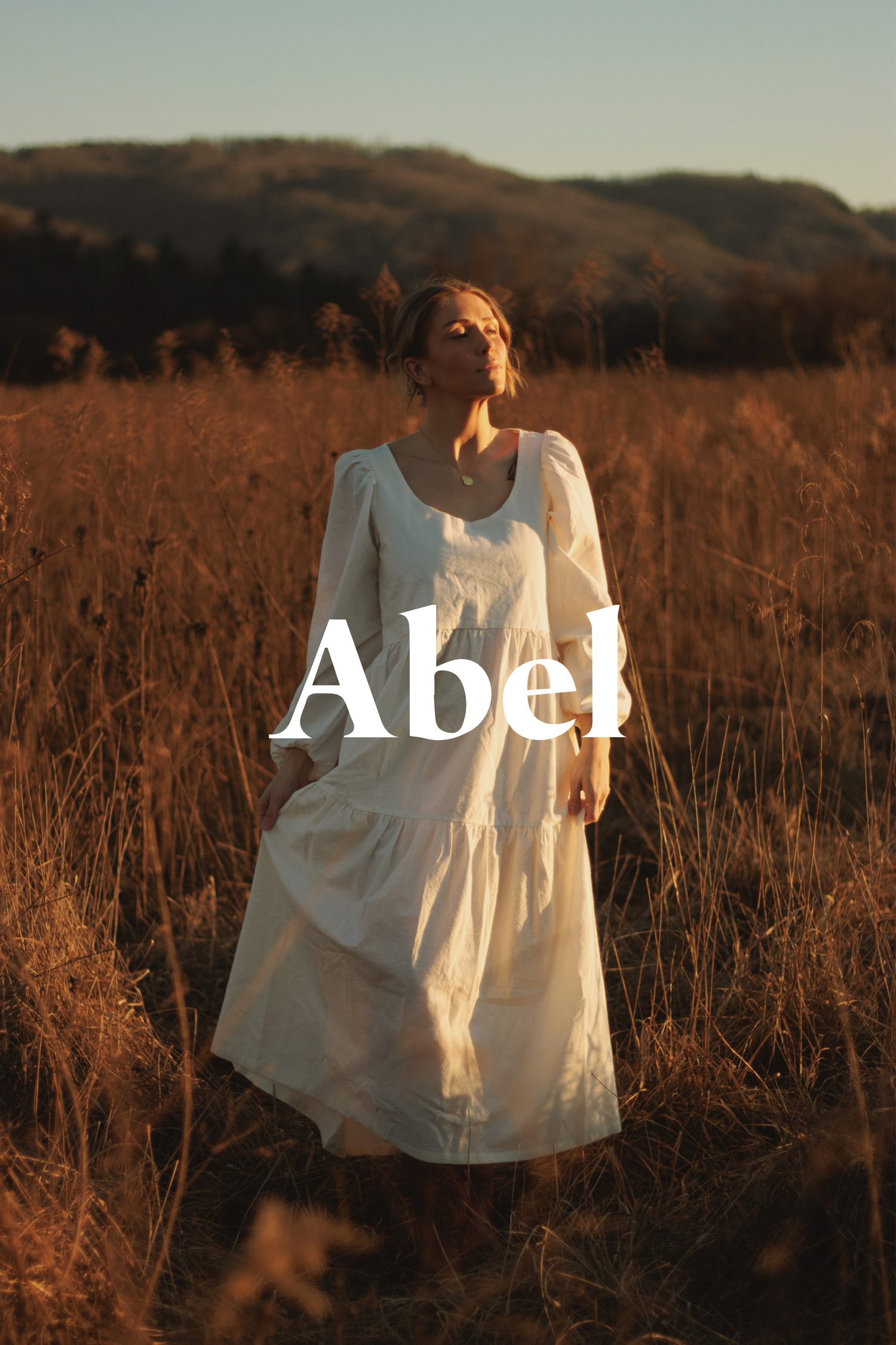 A figure in a flowing white dress stands in a golden field at sunset with rolling hills in the background.