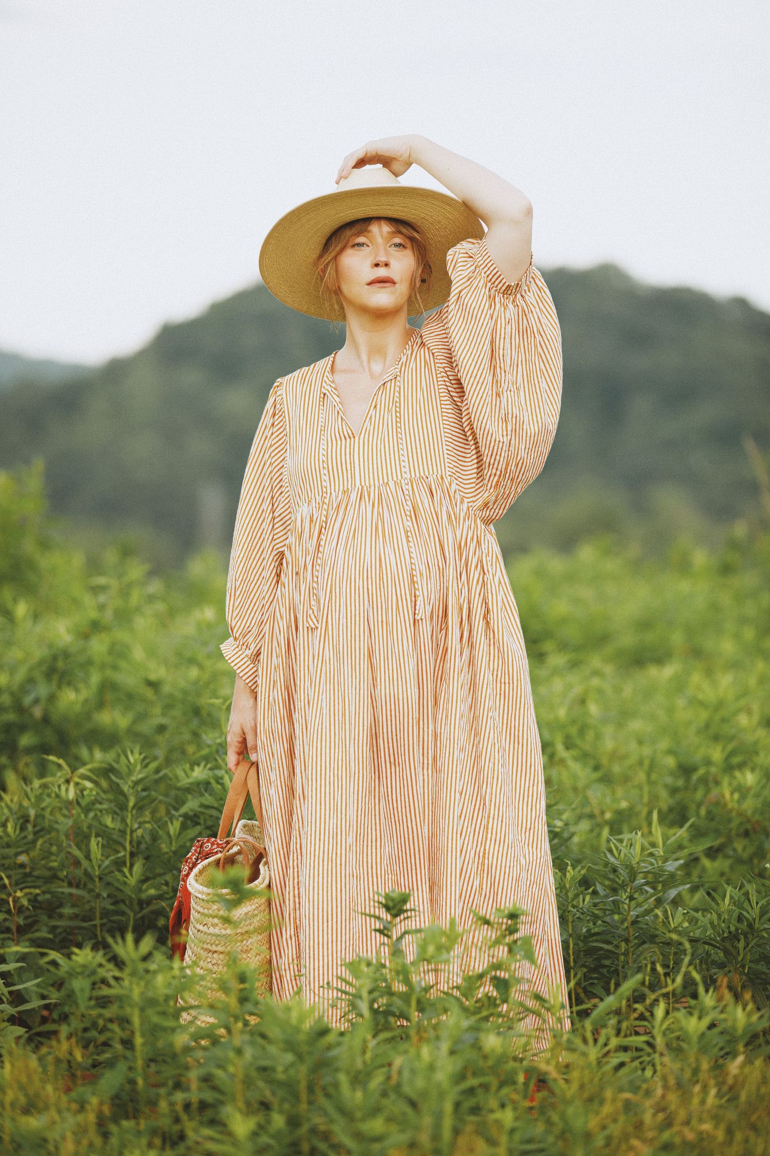 Woman in striped prairie dress and sun hat standing in wild meadow.