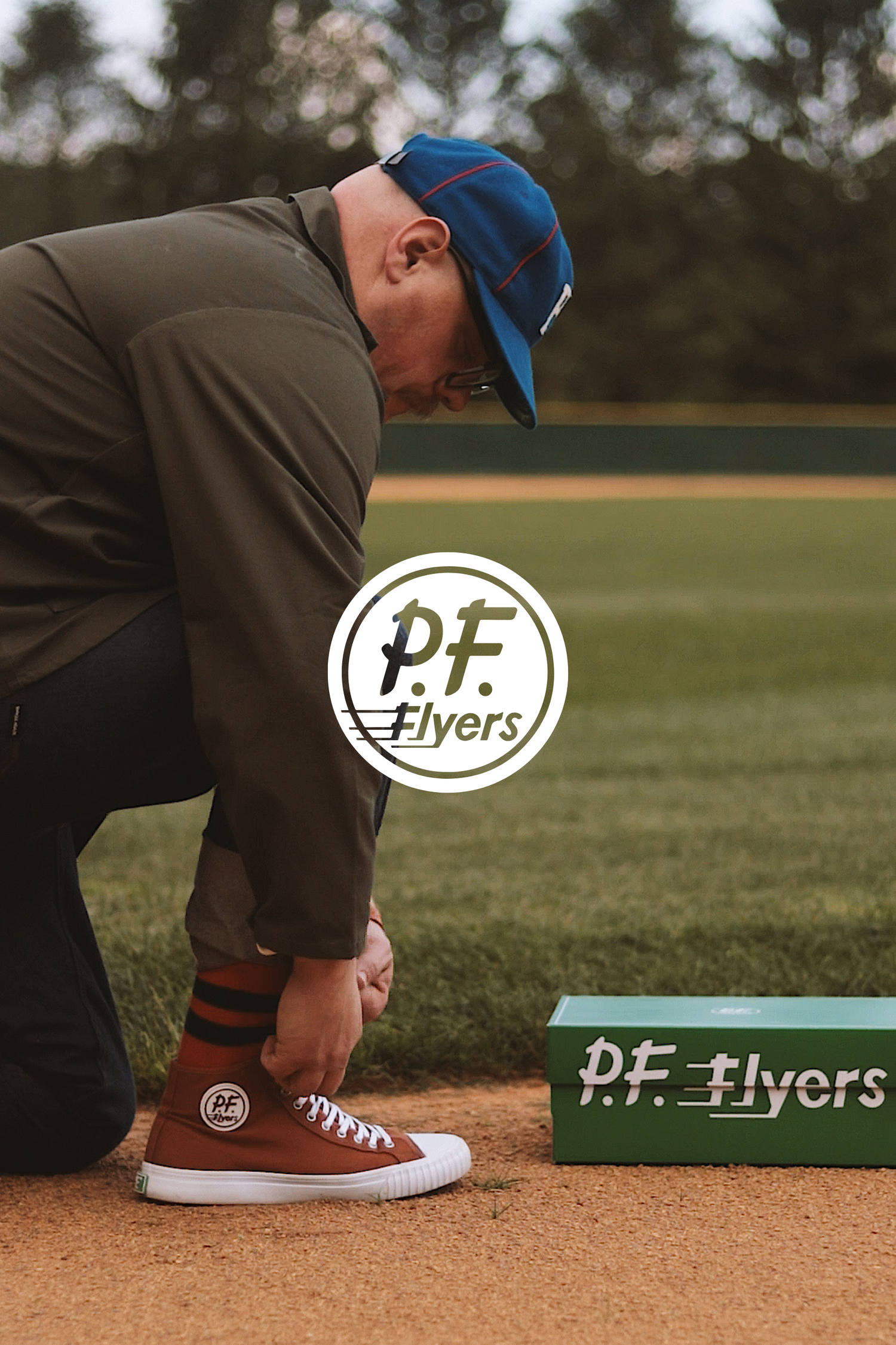 Person wearing a blue cap ties brown PF Flyers sneakers while standing on a baseball field with branding elements visible.