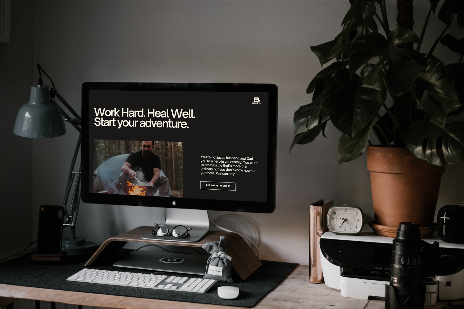A modern desk workspace setup with a dark monitor display, keyboard, potted plant, and desk lamp in moody lighting.
