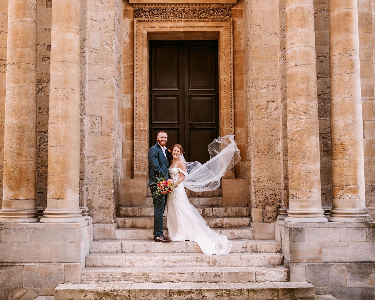 A wedding couple stands on stone steps of an ornate building with columns as the bride's veil flows dramatically in the wind.