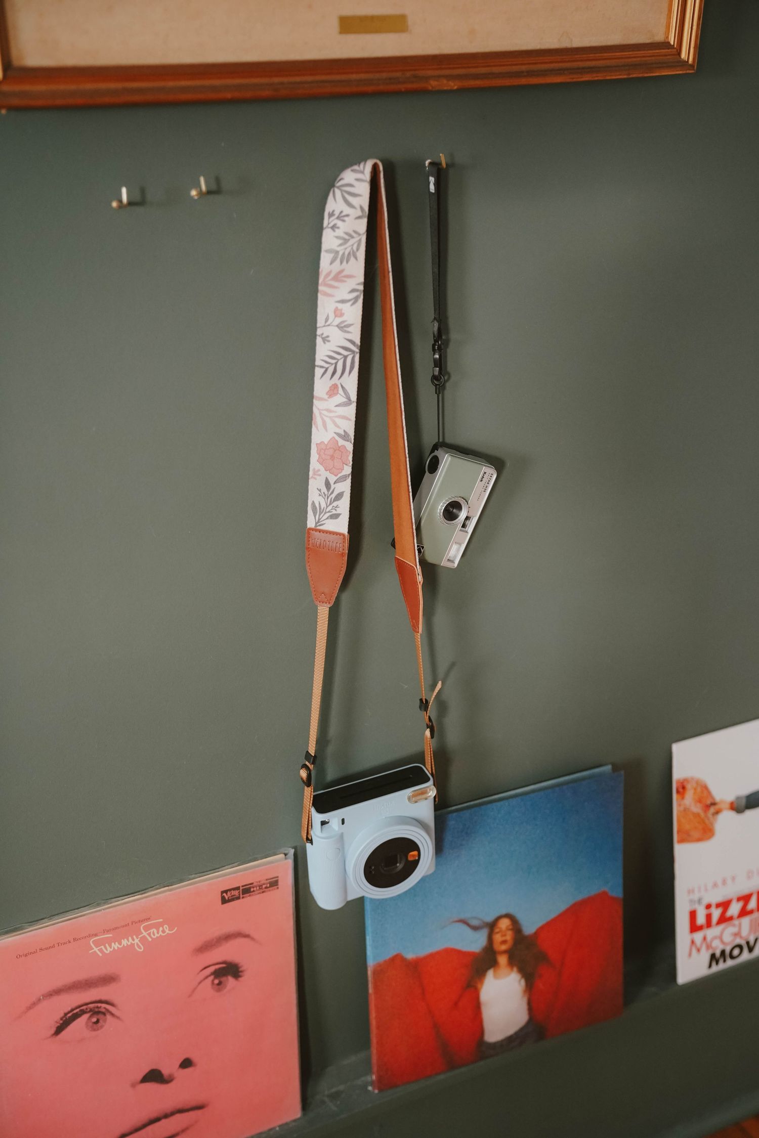 Camera with decorative strap hanging on sage green wall next to vinyl records and art prints.