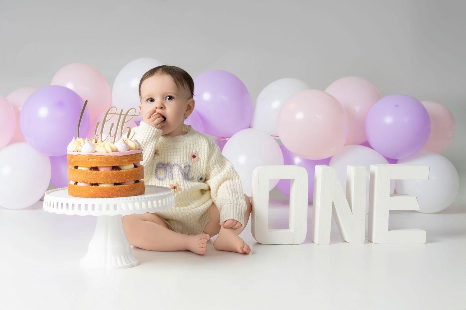 Baby sits with a smiling expression near a birthday cake with pink and purple balloons in the background.