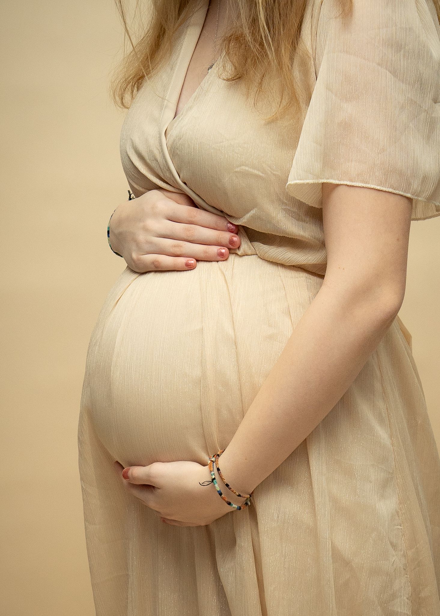 Close up of hands resting gently on a pregnant belly in a cream colored maternity dress with beige background.