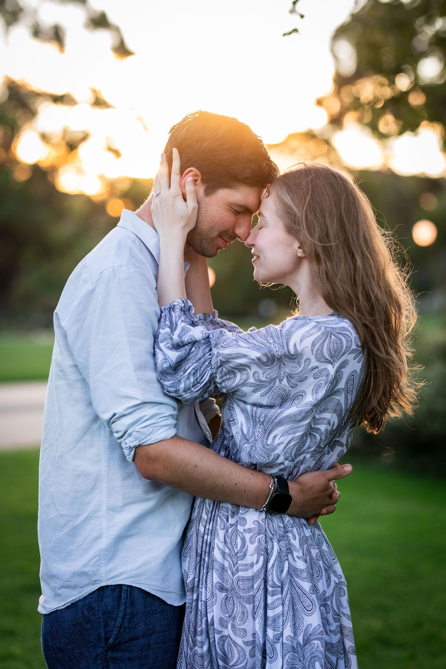 Engaged couple smiling and holding hands during their engagement photo session at Kings Park with greenery and city skyline in the backgroun