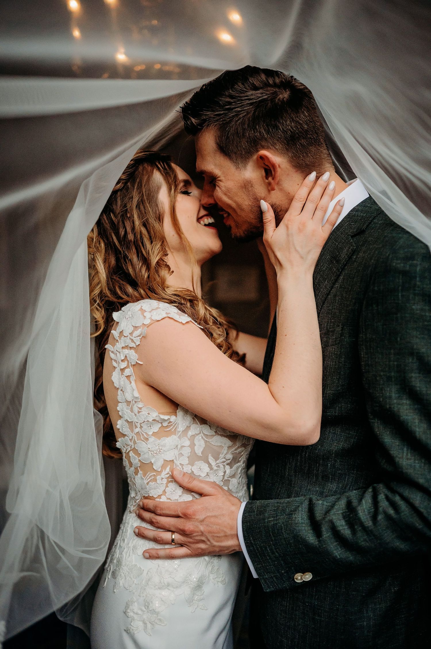 Intimate wedding portrait under flowing veil with couple sharing tender romantic moment in soft lighting.