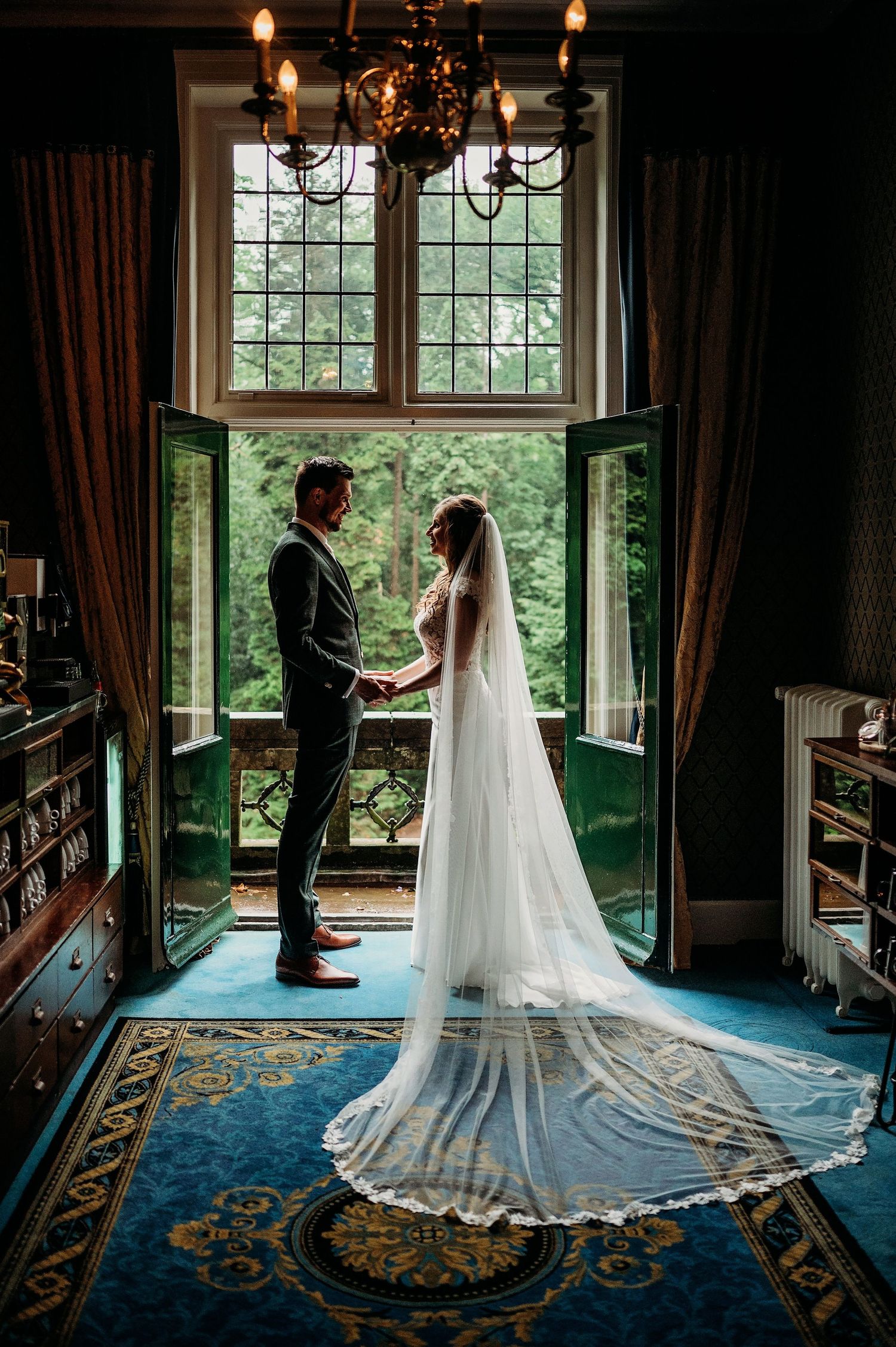 Two figures in wedding attire stand silhouetted by tall windows while a long lace veil drapes elegantly across a patterned blue rug.