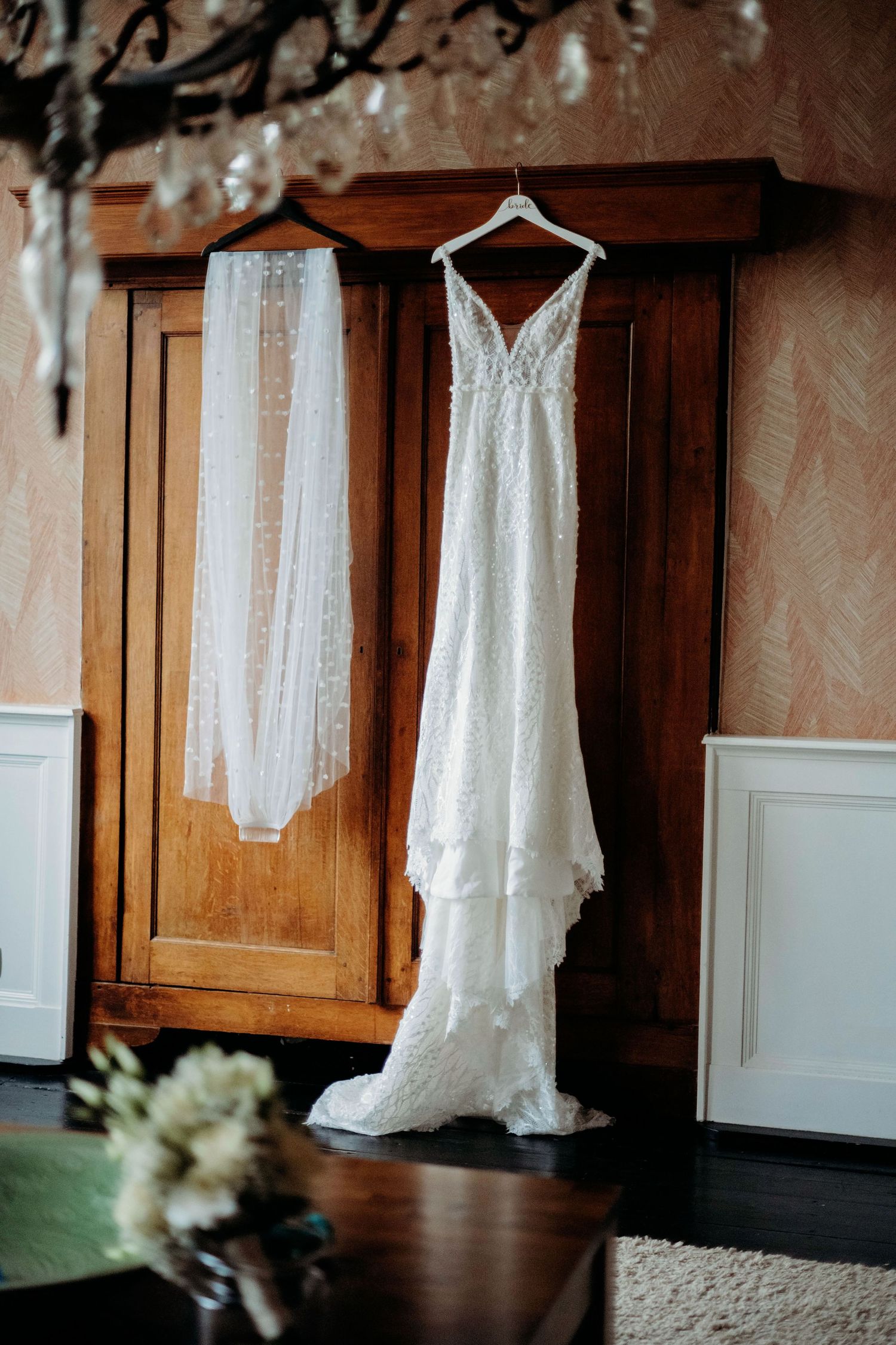 Elegant white lace wedding dress hanging on wooden wardrobe with veil alongside on rustic wooden background.