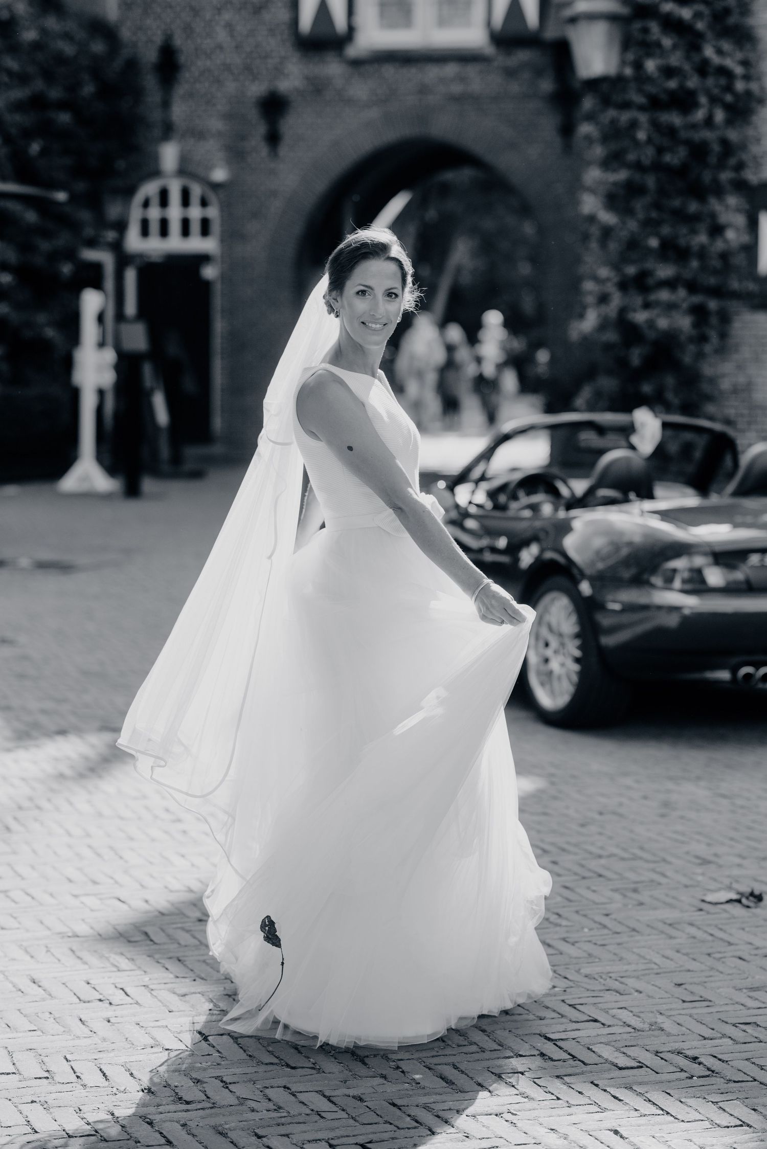 A bride in a flowing white wedding dress and veil twirls on a brick courtyard with a castle-like entrance in the background.