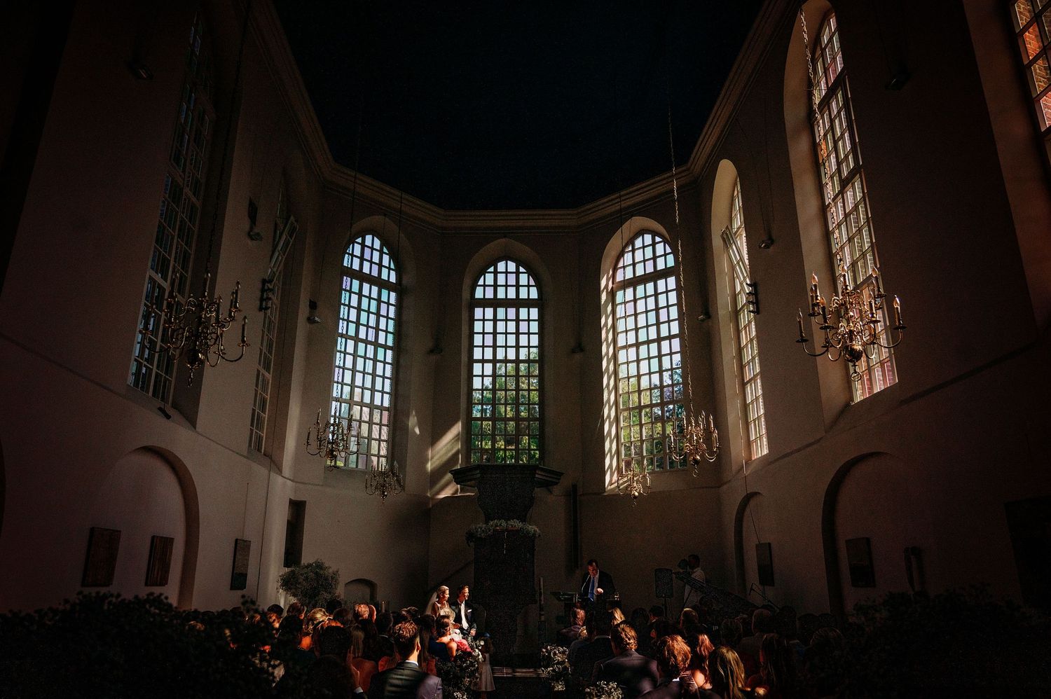 A dark church interior with arched windows illuminating performers and audience members during an evening concert.