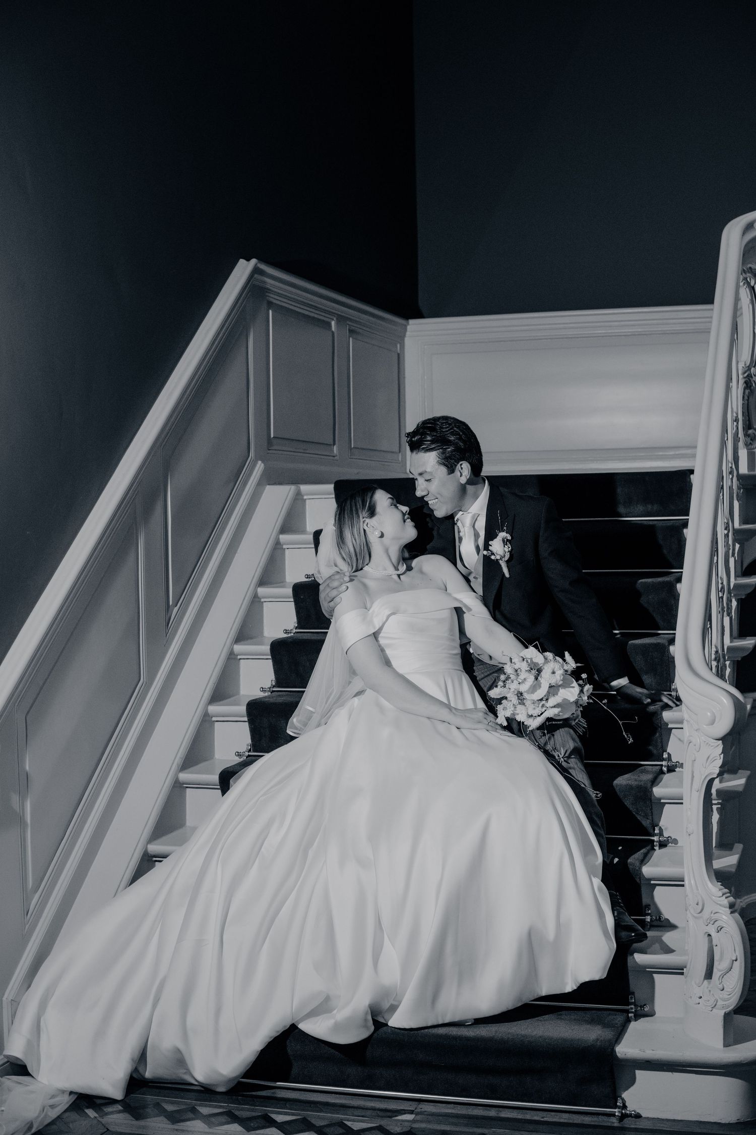 A bride in a flowing white ballgown and groom share a romantic moment on an elegant staircase at their wedding.