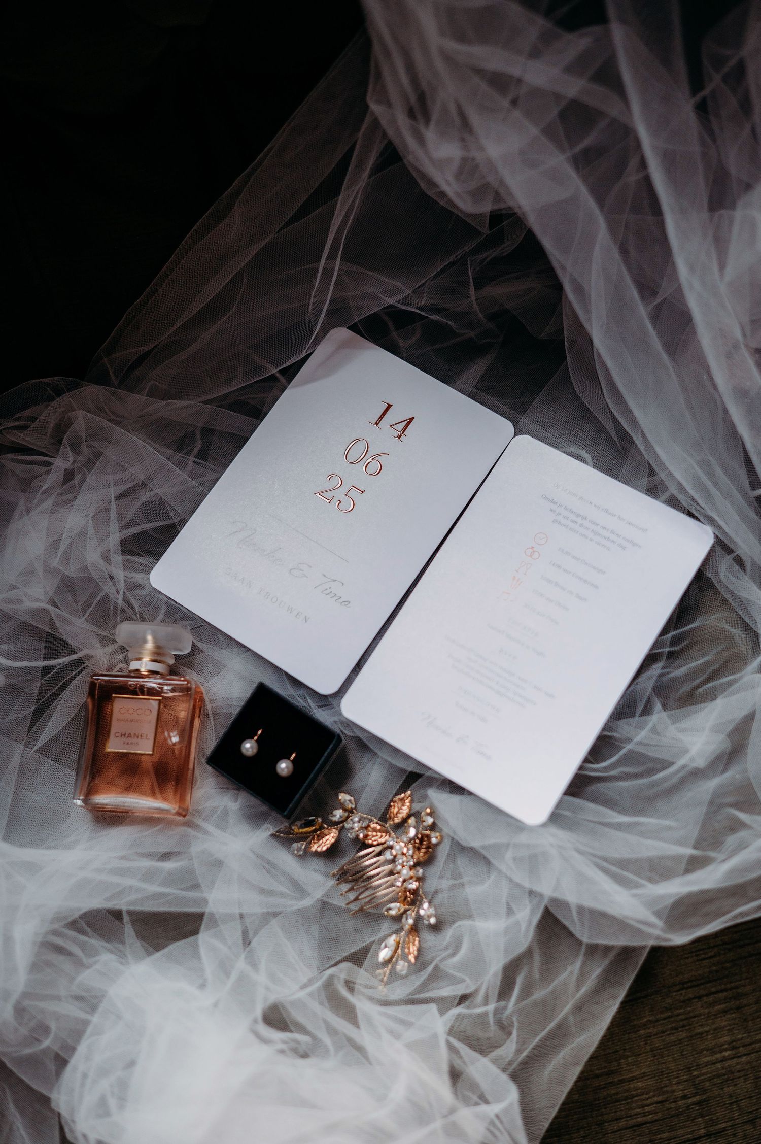 Wedding details including perfume bottle, invitation cards, jewelry and delicate tulle veil arranged on dark surface.