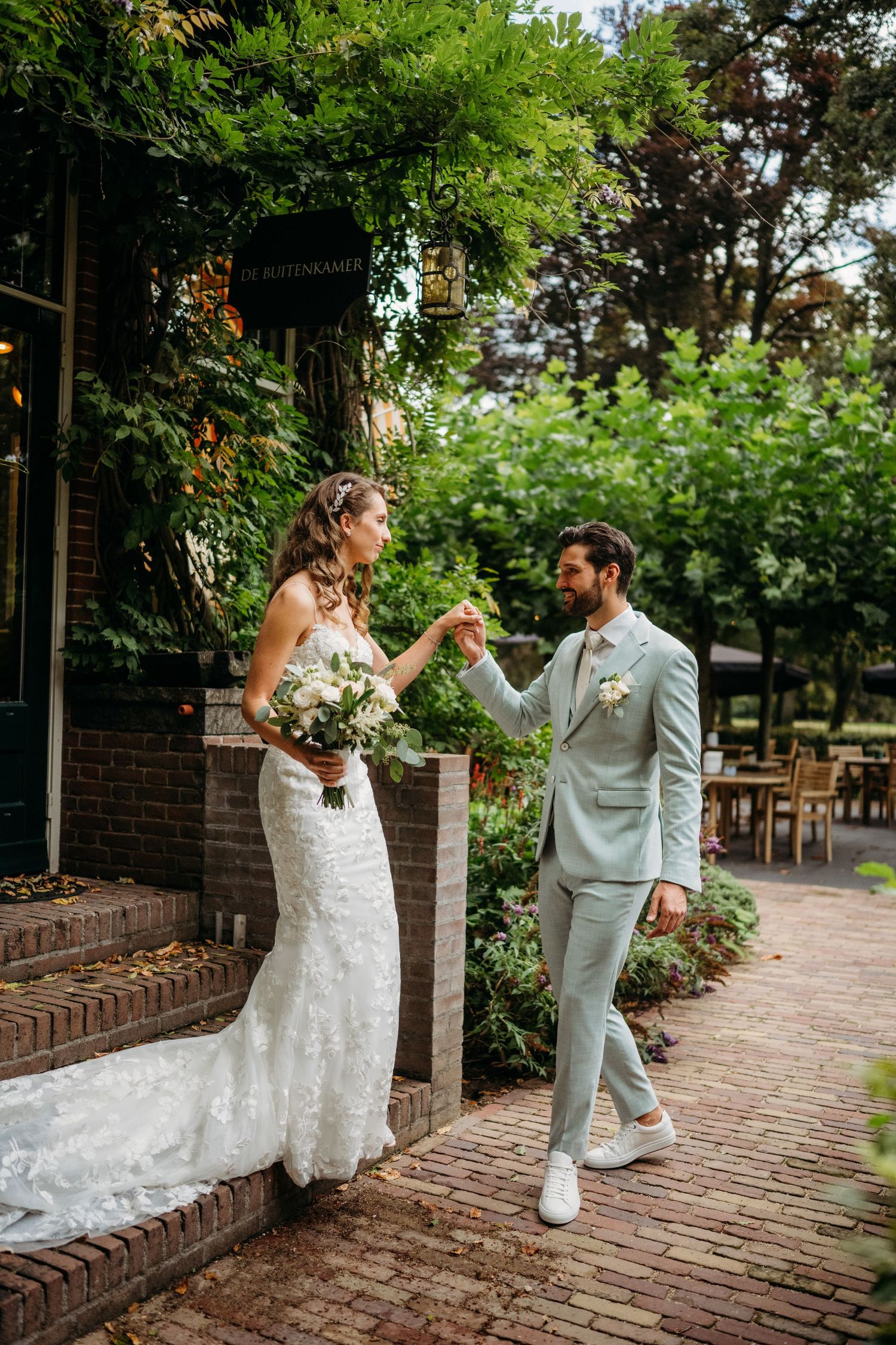 A couple in wedding attire share a romantic moment on a brick pathway surrounded by lush greenery and trees.