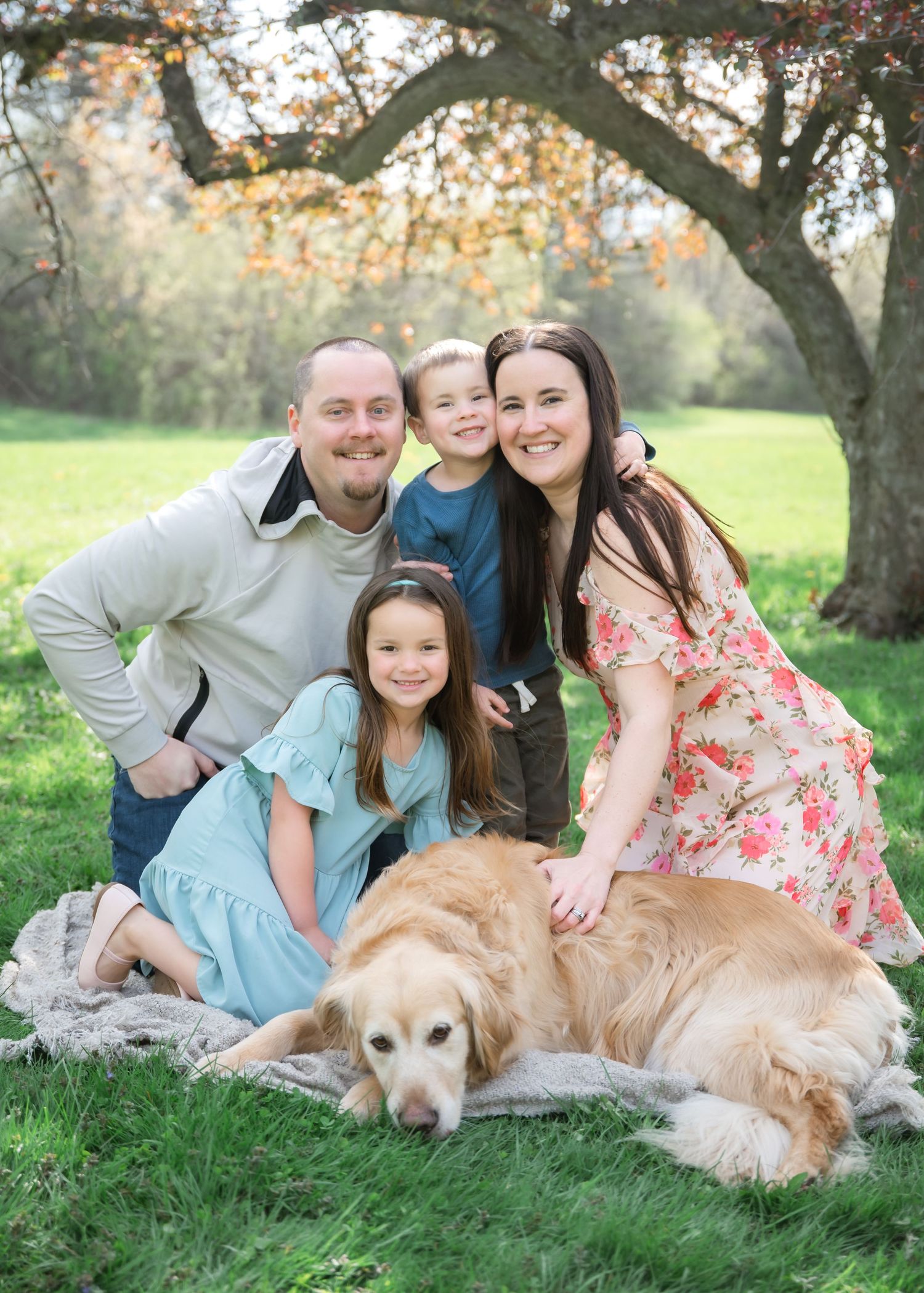 A Golden Retriever rests on green grass beneath an autumn tree while a family gathers around for an outdoor portrait.