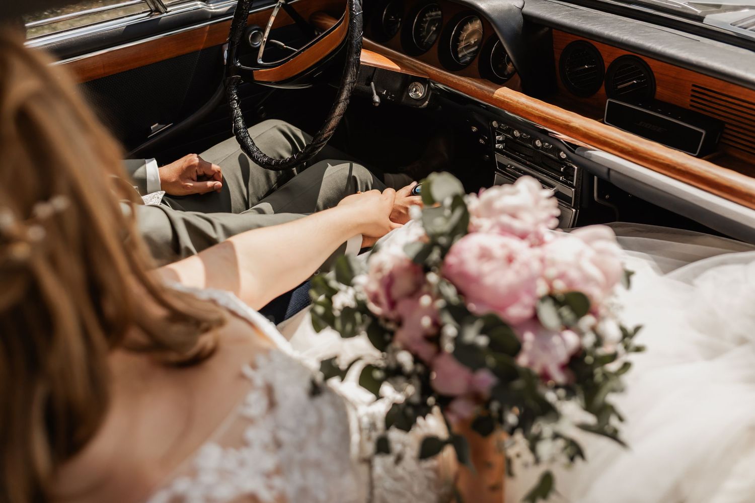 Pink peonies and wedding dress visible in vintage car interior with wood grain dashboard and steering wheel.