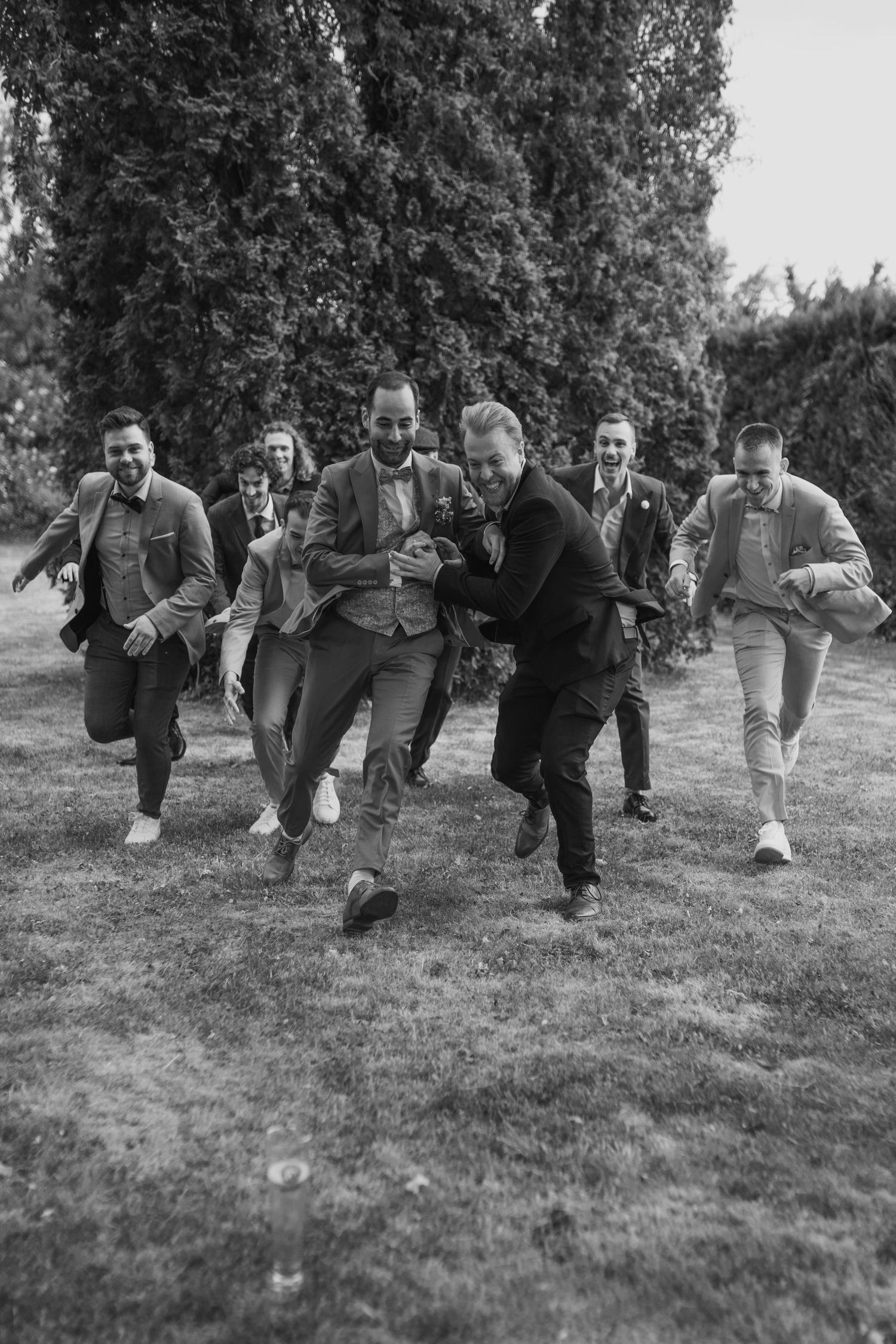 Group of groomsmen running playfully on grass in black and white photography.