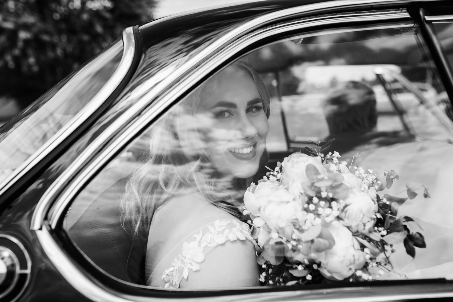 Elegant bride with bouquet smiling through vintage car window in black and white.