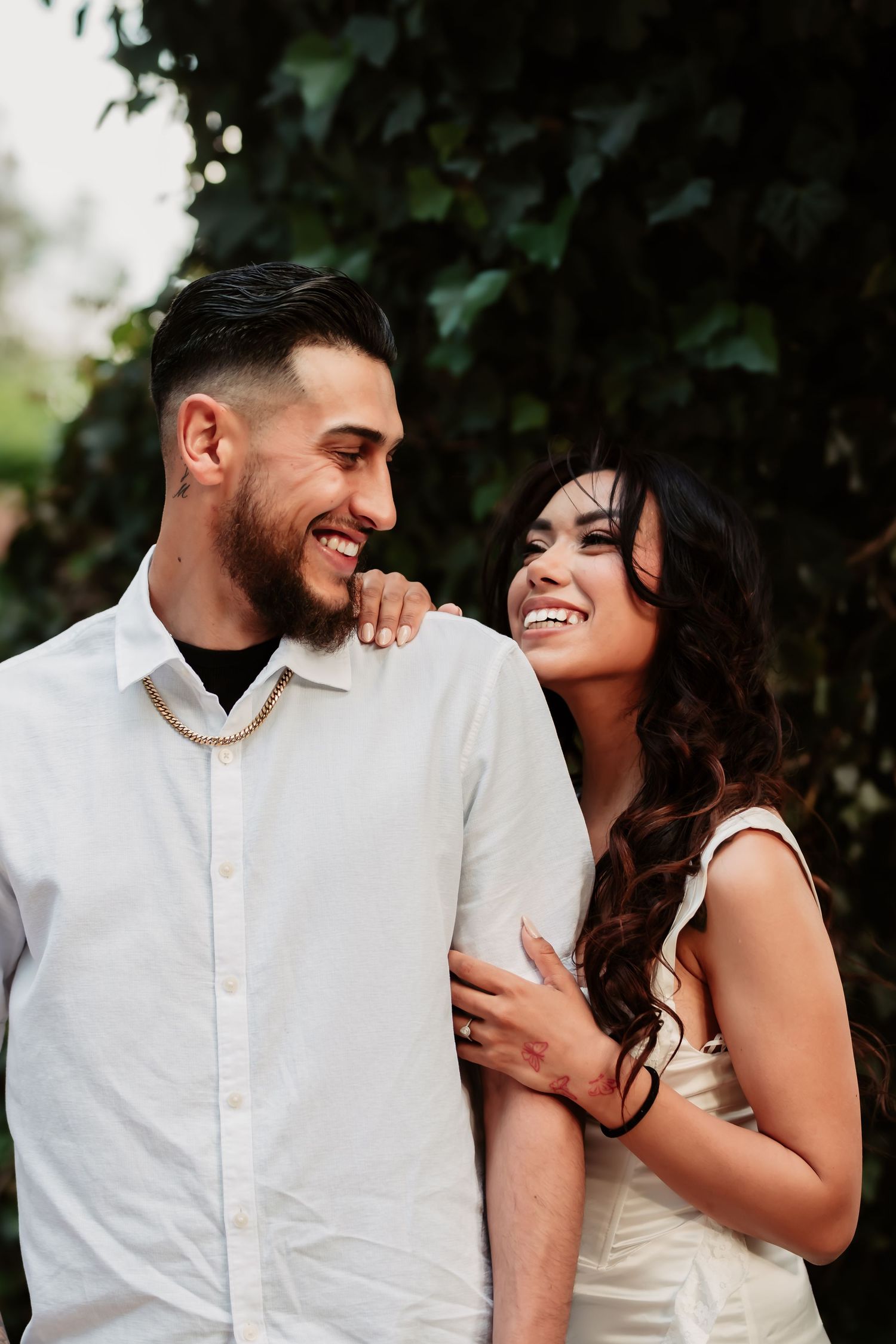 A romantic couple sharing a joyful laugh together outdoors with lush greenery in the background.