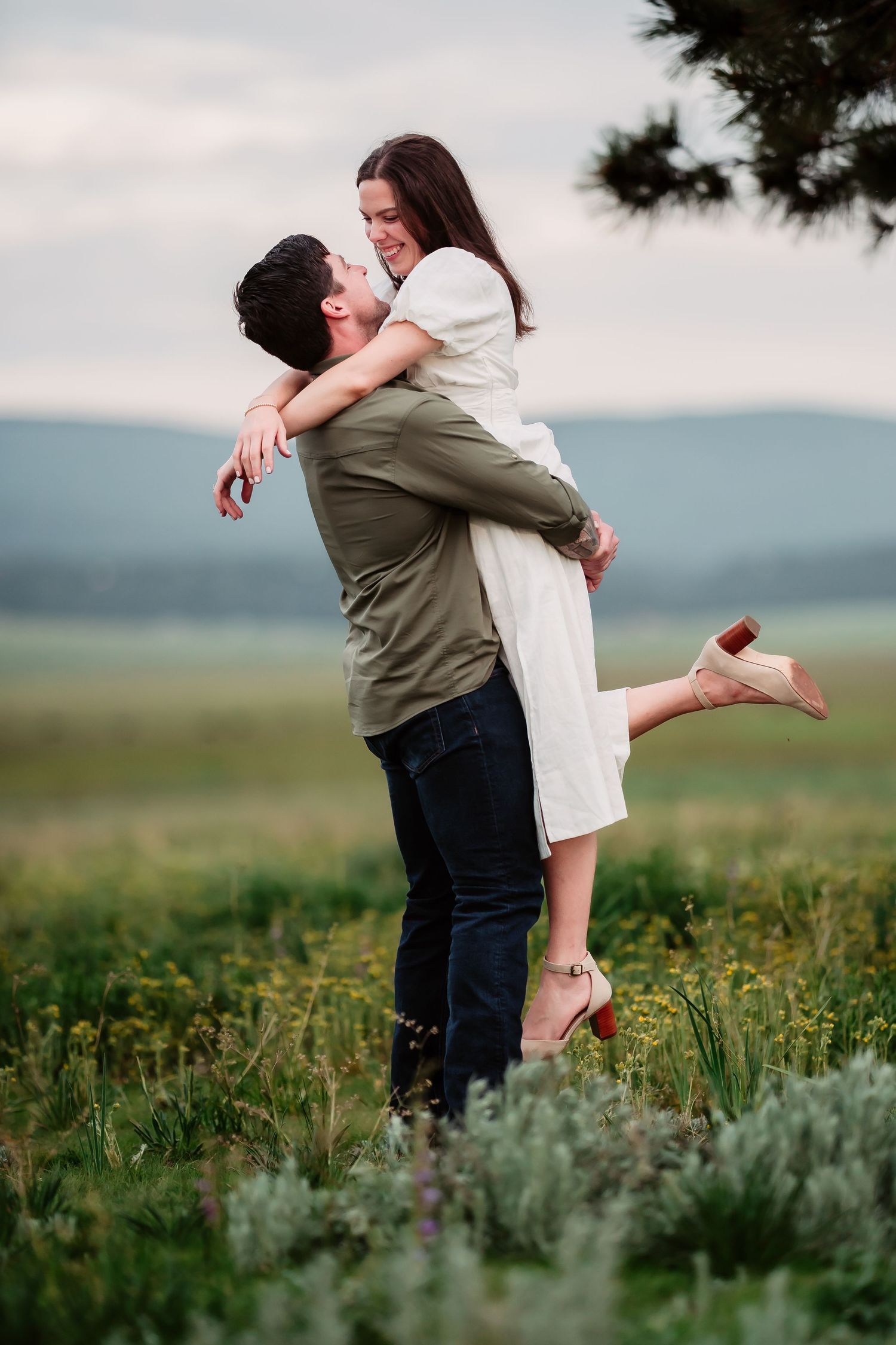 A romantic couple embracing in a field of wildflowers during sunset with mountains in the background.