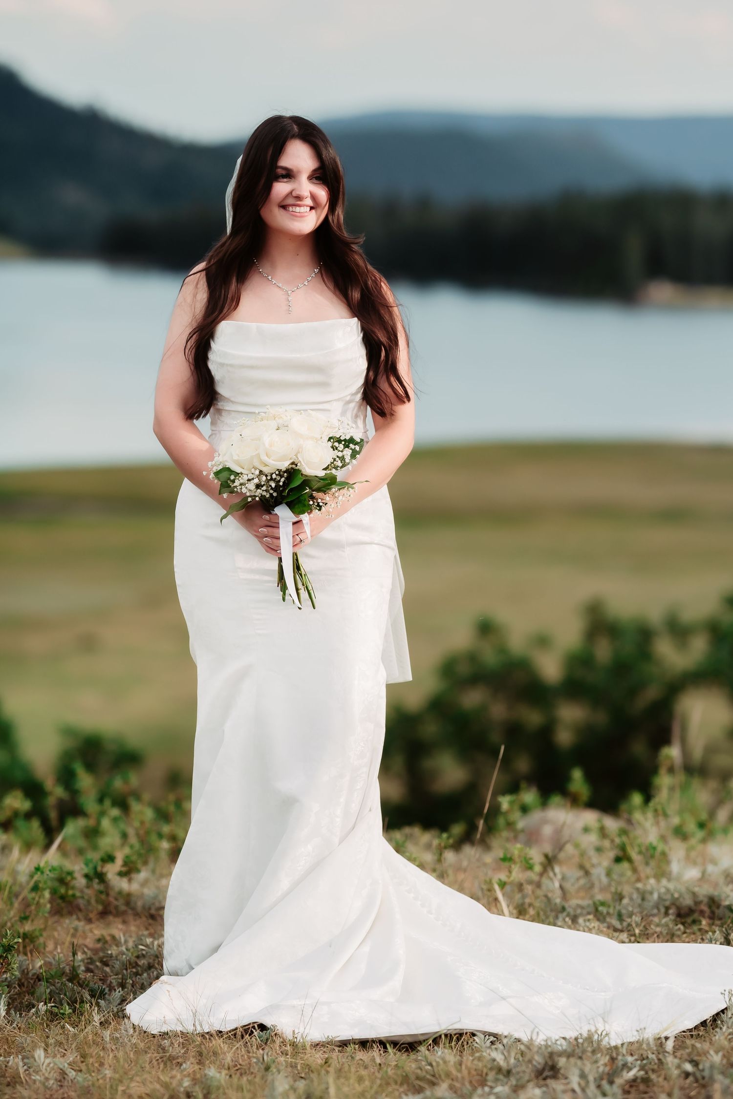 A bride in a strapless wedding gown holds a small bouquet while posing near a scenic lake.