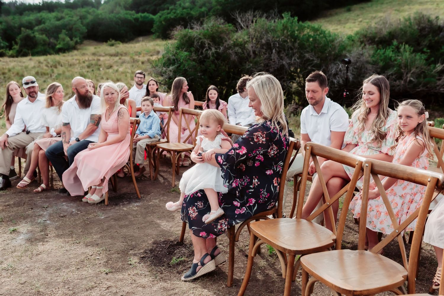 Wedding guests in light summer attire seated on wooden chairs at outdoor mountain ceremony.